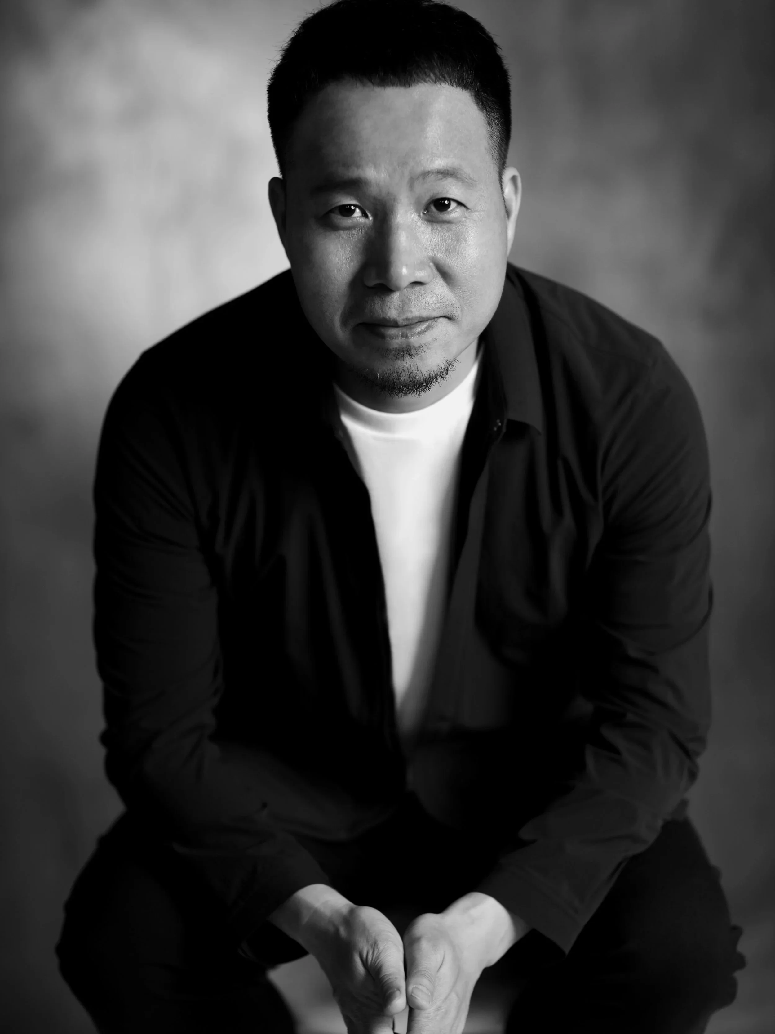 A black-and-white portrait of a man with short hair and a slight beard, sitting with his hands clasped, looking directly at the camera.