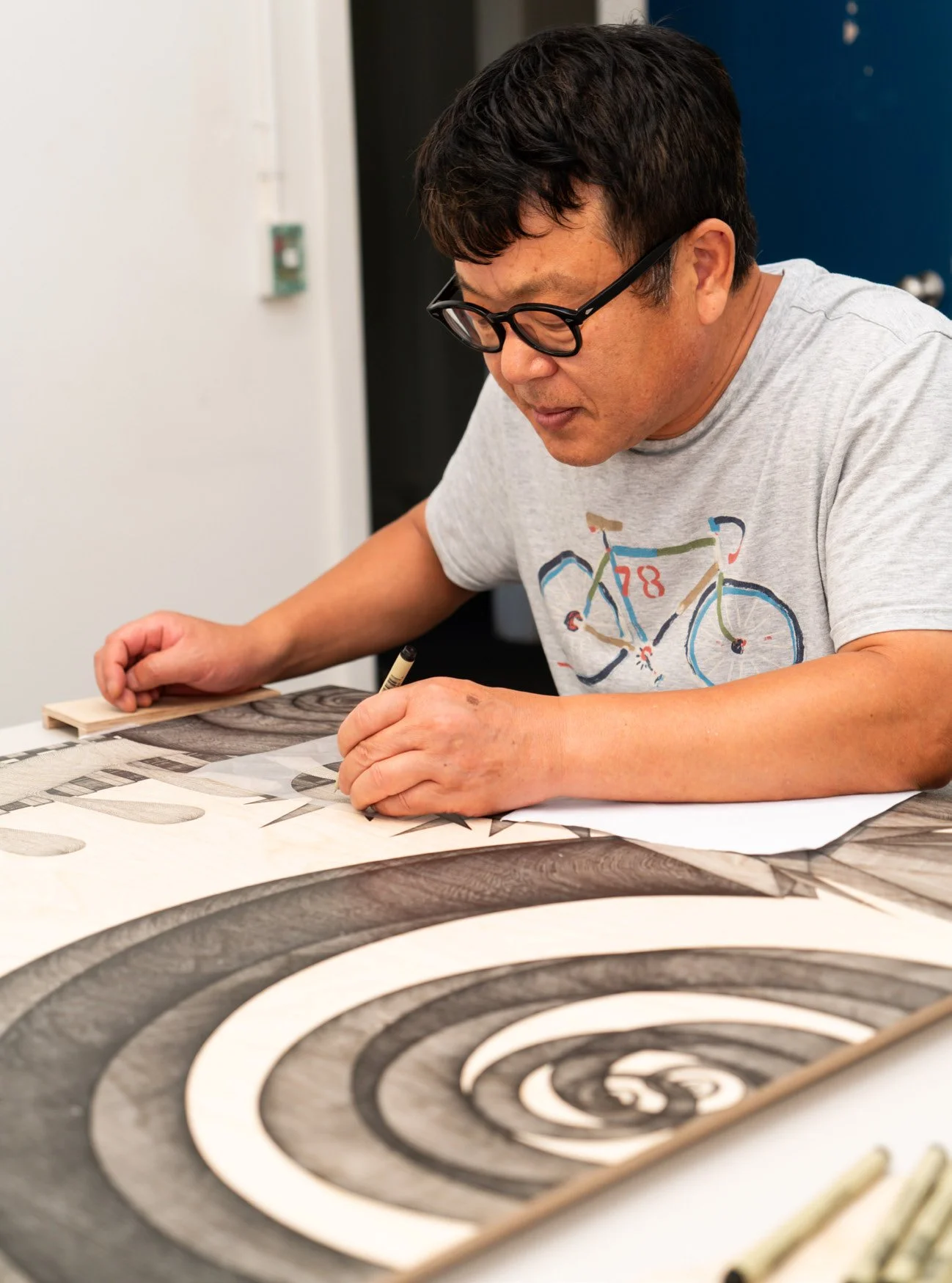 A man wearing glasses working on a detailed black and white spiraling pattern on paper, sitting at a table.