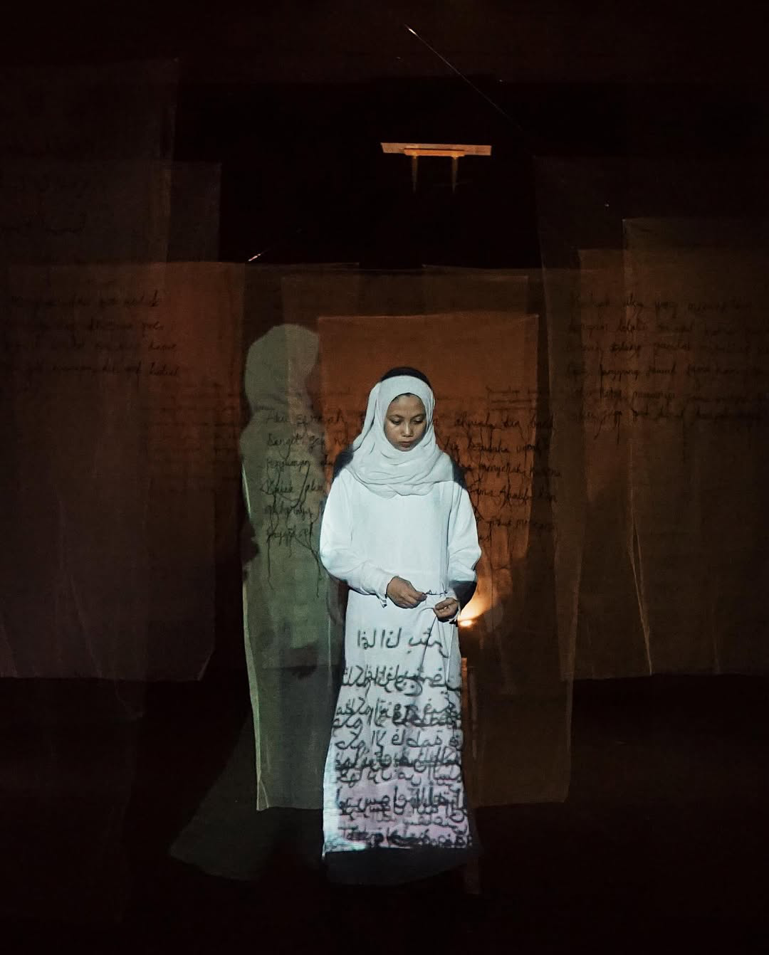 A young woman dressed in white with a headscarf stands in a dark room, illuminated by projected handwritten Arabic script on transparent sheets around her.