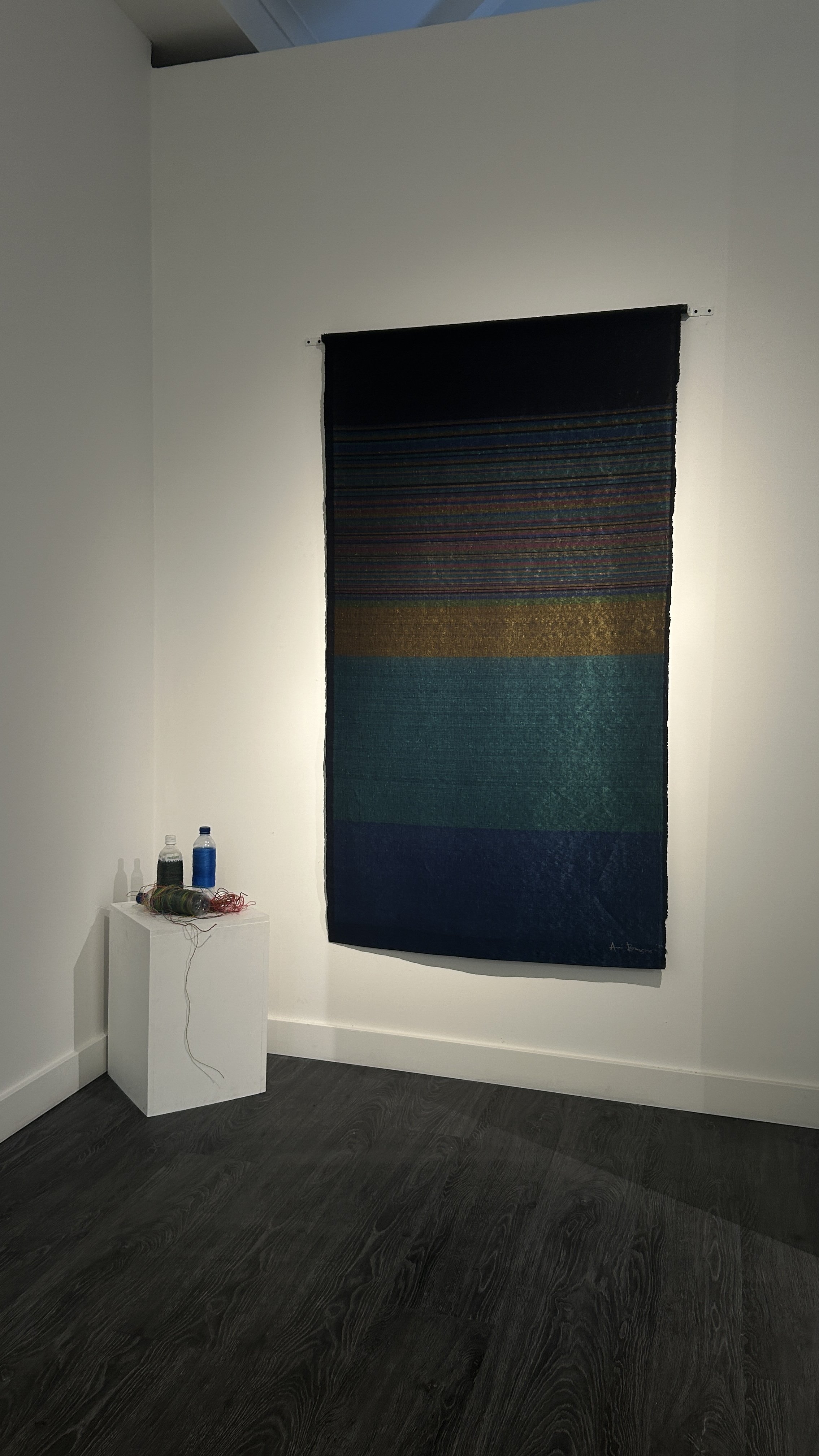 A colorful textile wall hanging displayed on a white gallery wall, with a small white pedestal below holding various spools of thread and yarn, in a modern art gallery setting.