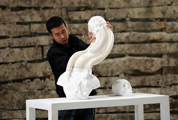 An artist working on a large white sculpture of a human head with an elongated, swirling neck. Another smaller skull sculpture is on the table beside him. The background consists of wooden planks.