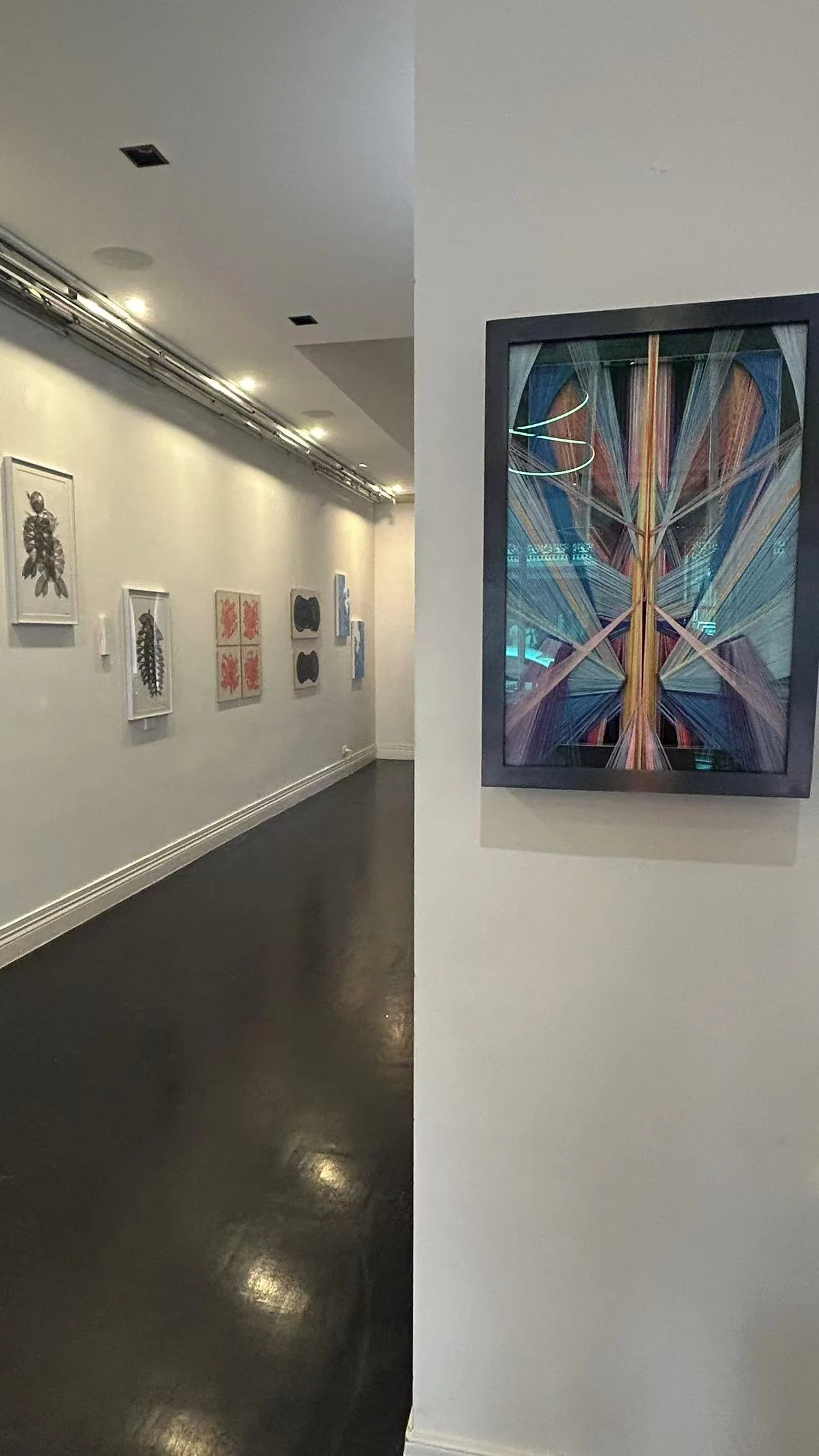 An art gallery with white walls displaying various framed artworks. The foreground features a colorful, abstract framed piece on the right, while the background has a wall with multiple black and white illustrations and abstract paintings. The galler