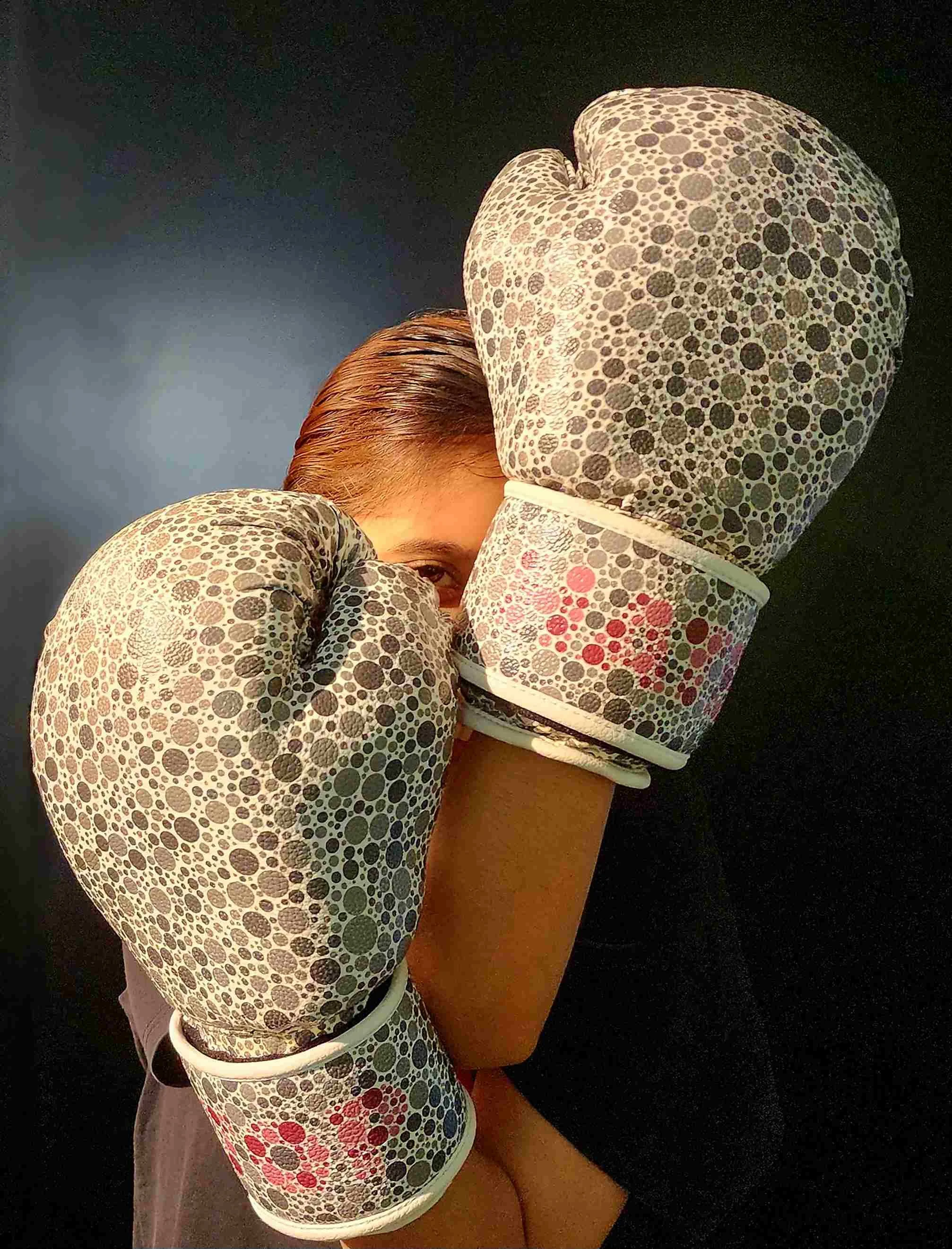A person wearing patterned oven mitts with black, gray, and red circles. Their face is partially visible, with one eye peeking out from behind the mitts. The person is holding a large rectangular object, possibly a wrapped gift or box, with a dark ba