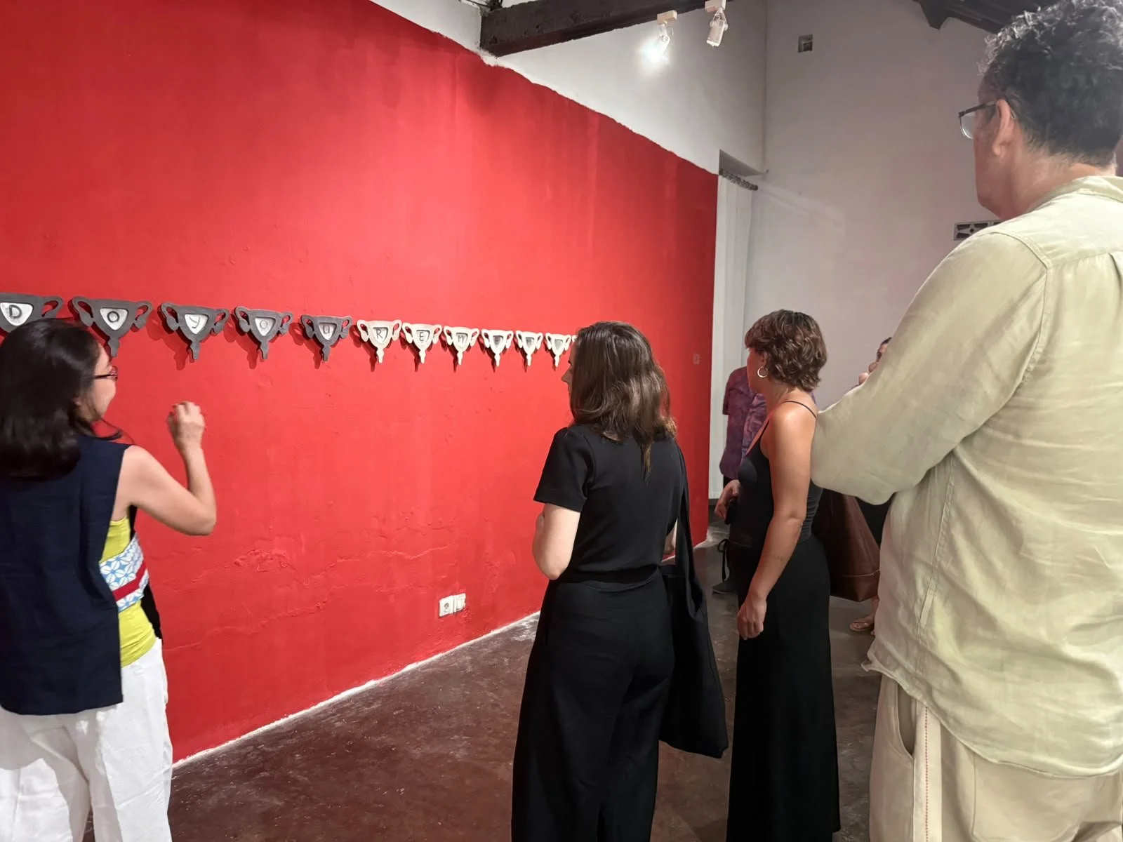 A group of people observing an art installation of a string of colorful, abstract triangular objects displayed on a red wall.