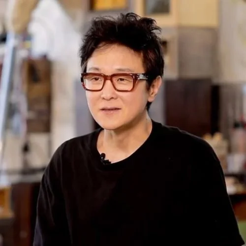 A woman with short black hair and glasses sitting indoors in a casual setting.