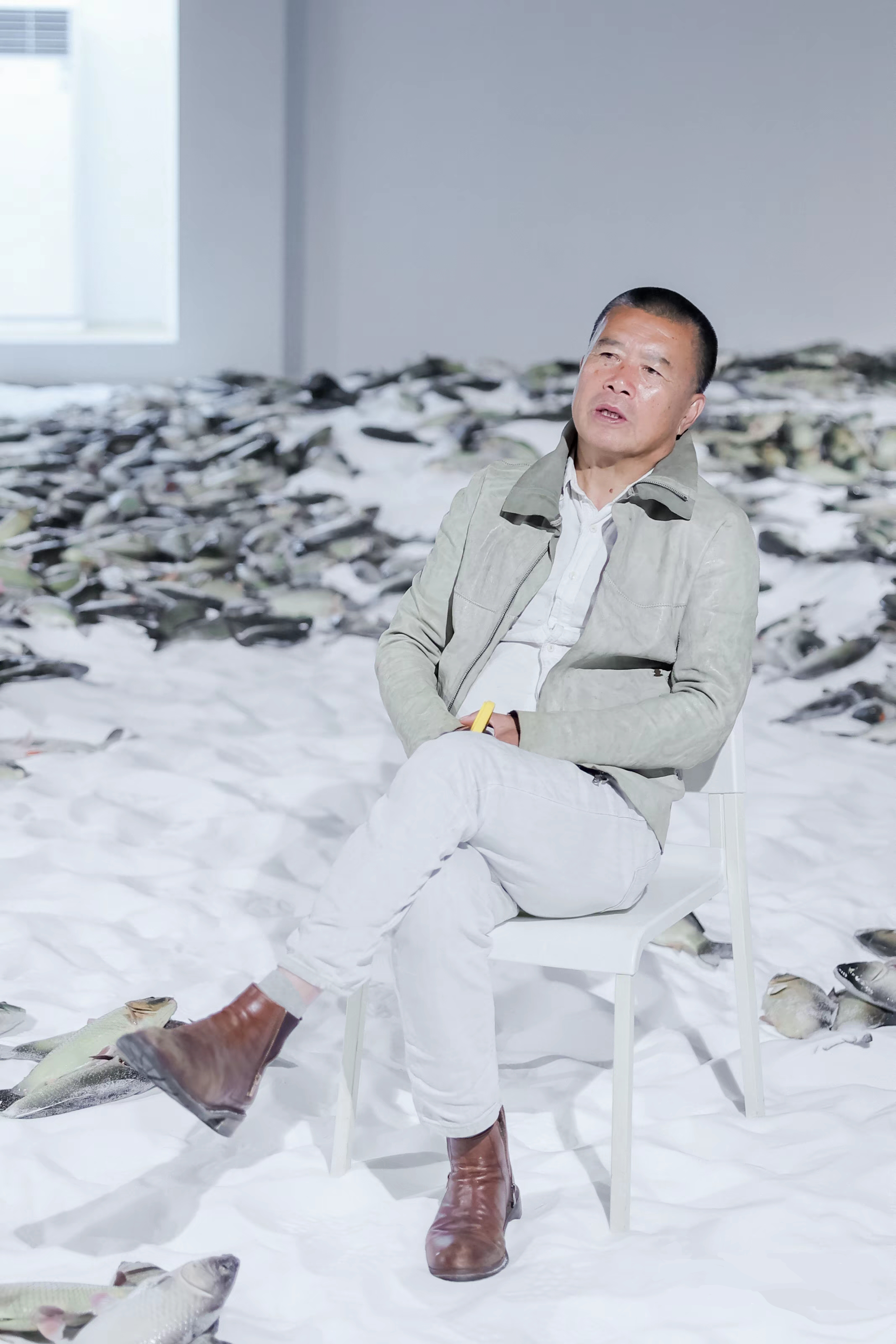 A man wearing a beige jacket and white pants sitting on a white chair in an art installation or exhibit filled with fish scattered on the floor and piled on a table, with a white wall and window in the background.