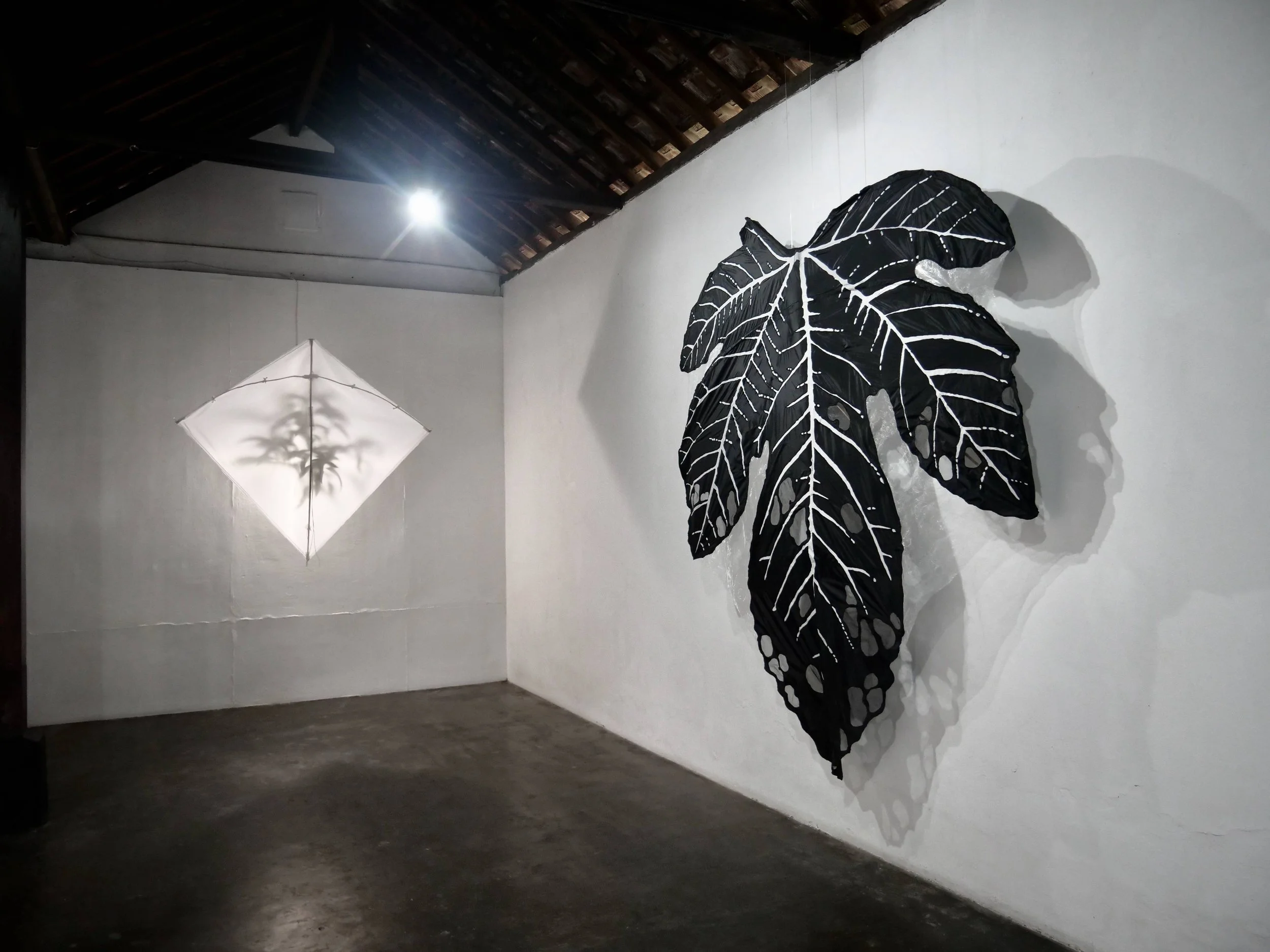Indoor art installation featuring black and white butterfly and leaf design on fabric, with a white umbrella-shaped light fixture on the wall, in a gallery setting with white walls and wooden ceiling.