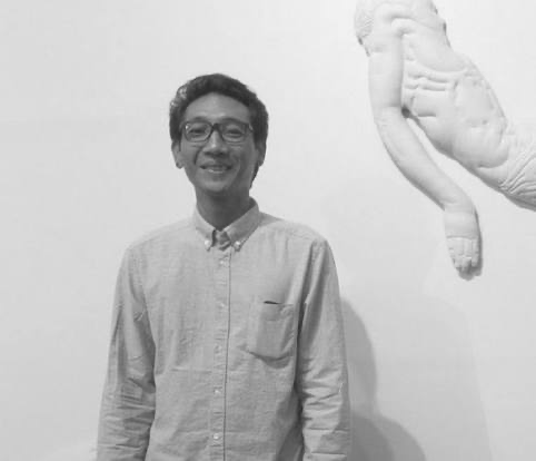 A man smiling and wearing glasses, standing next to a large sculpture of an insect's abdomen on a plain white wall.