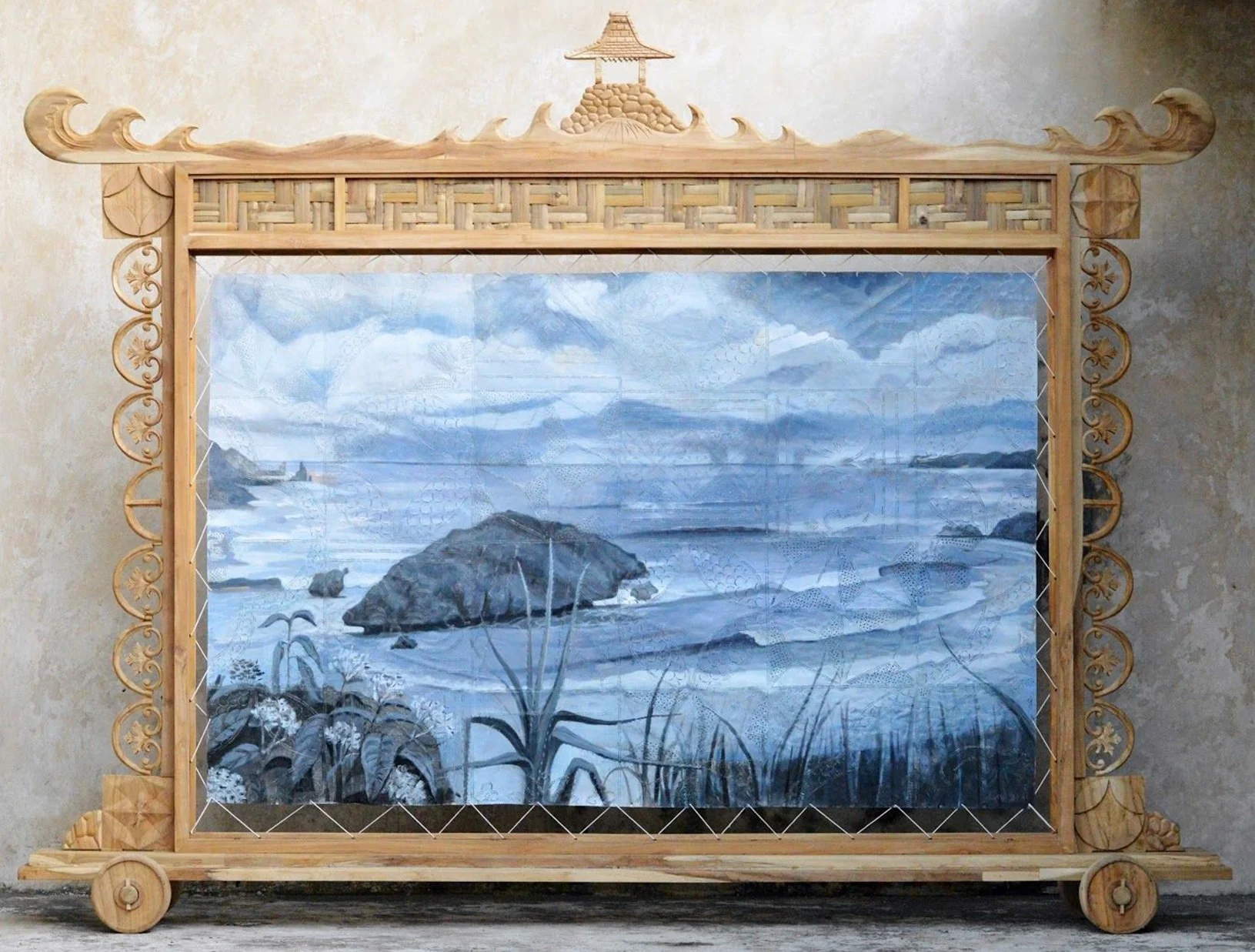 A wooden framed mirror with intricately carved sides and top, featuring a small pagoda structure at the top center. The mirror reflects a landscape painting of a rocky shoreline with a large rock in the water, mountains in the distance, and a cloudy 
