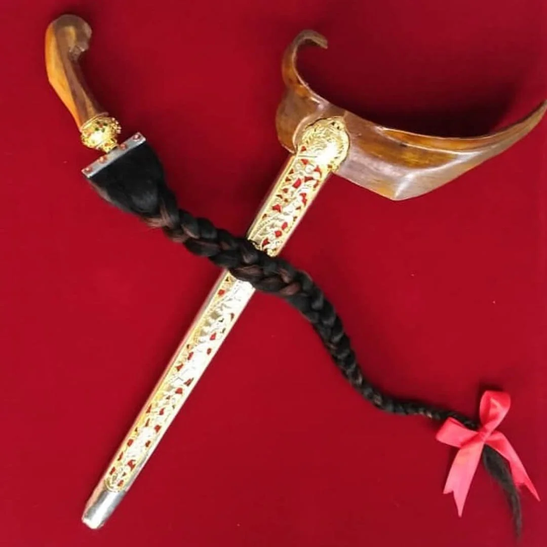 Decorative dagger with a curved blade, gold detailing, black handle with gold embellishments, and a black braided hair with a pink bow attached, set against a red background.