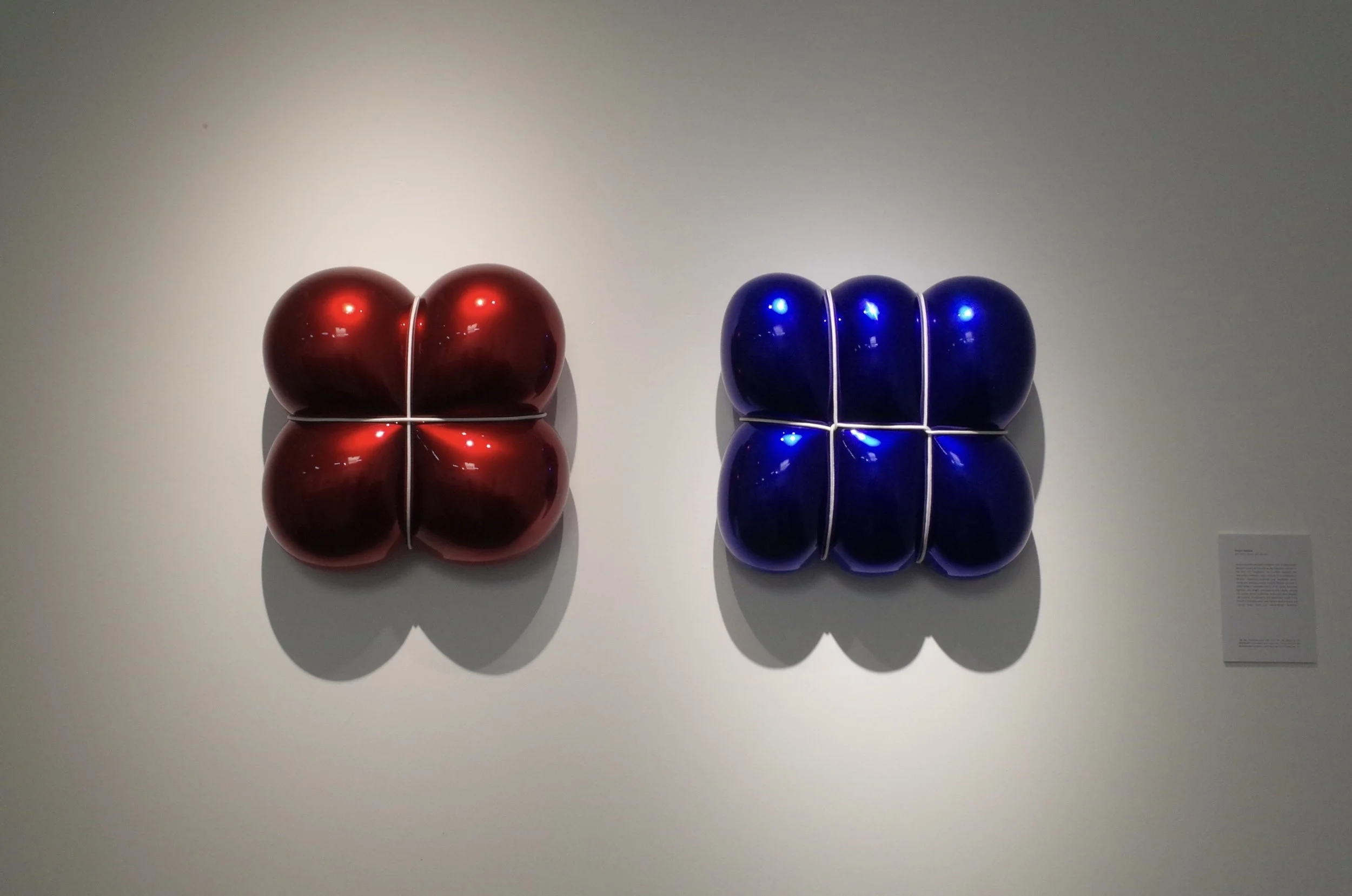 Two sets of balloon sculptures, one red and one blue, arranged in a square shape on a white wall, casting shadows.