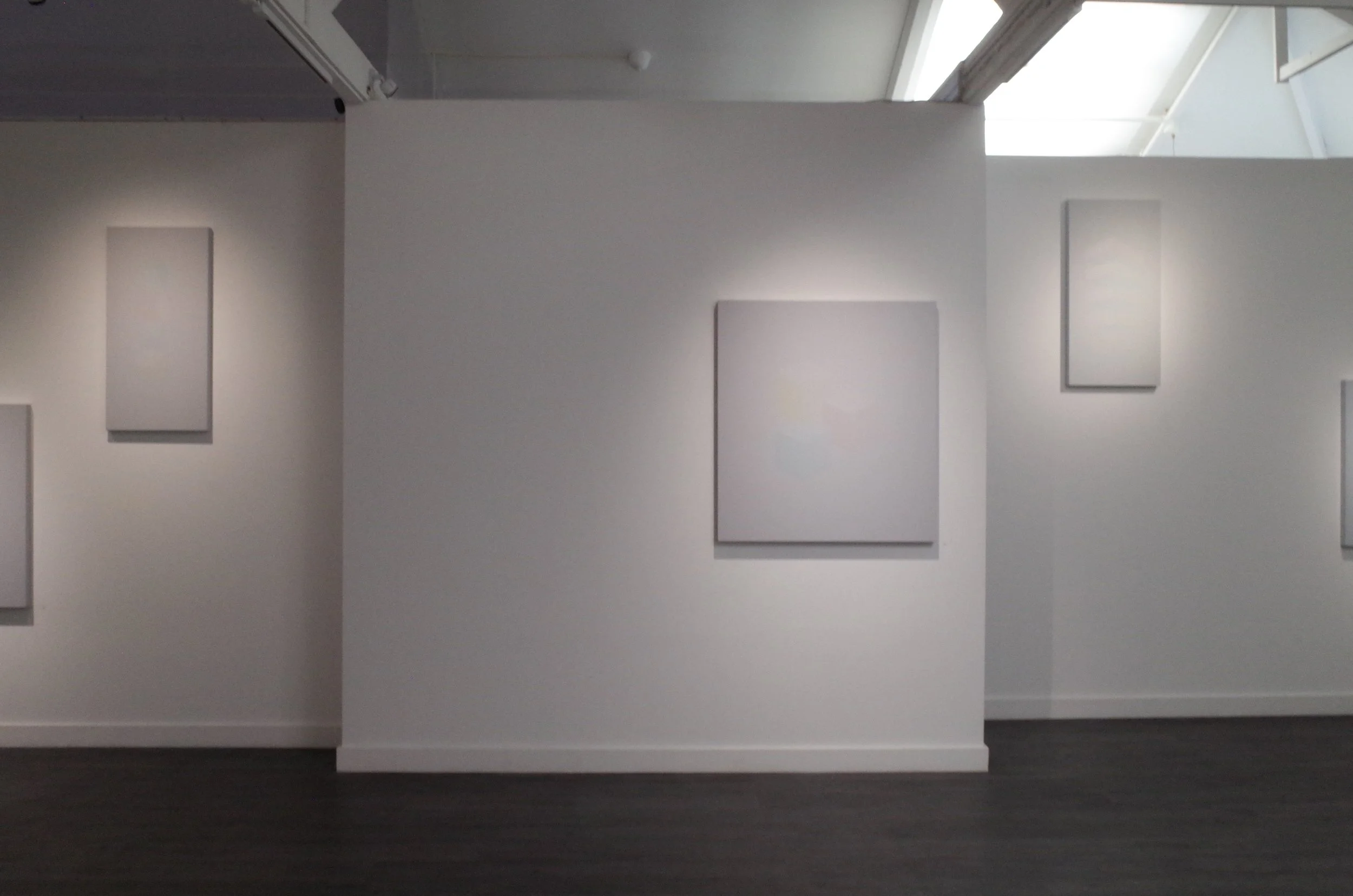 A white gallery wall displaying six blank canvases of various sizes, with three on the left and three on the right, under a sloped ceiling with skylights, in an art gallery or museum setting.
