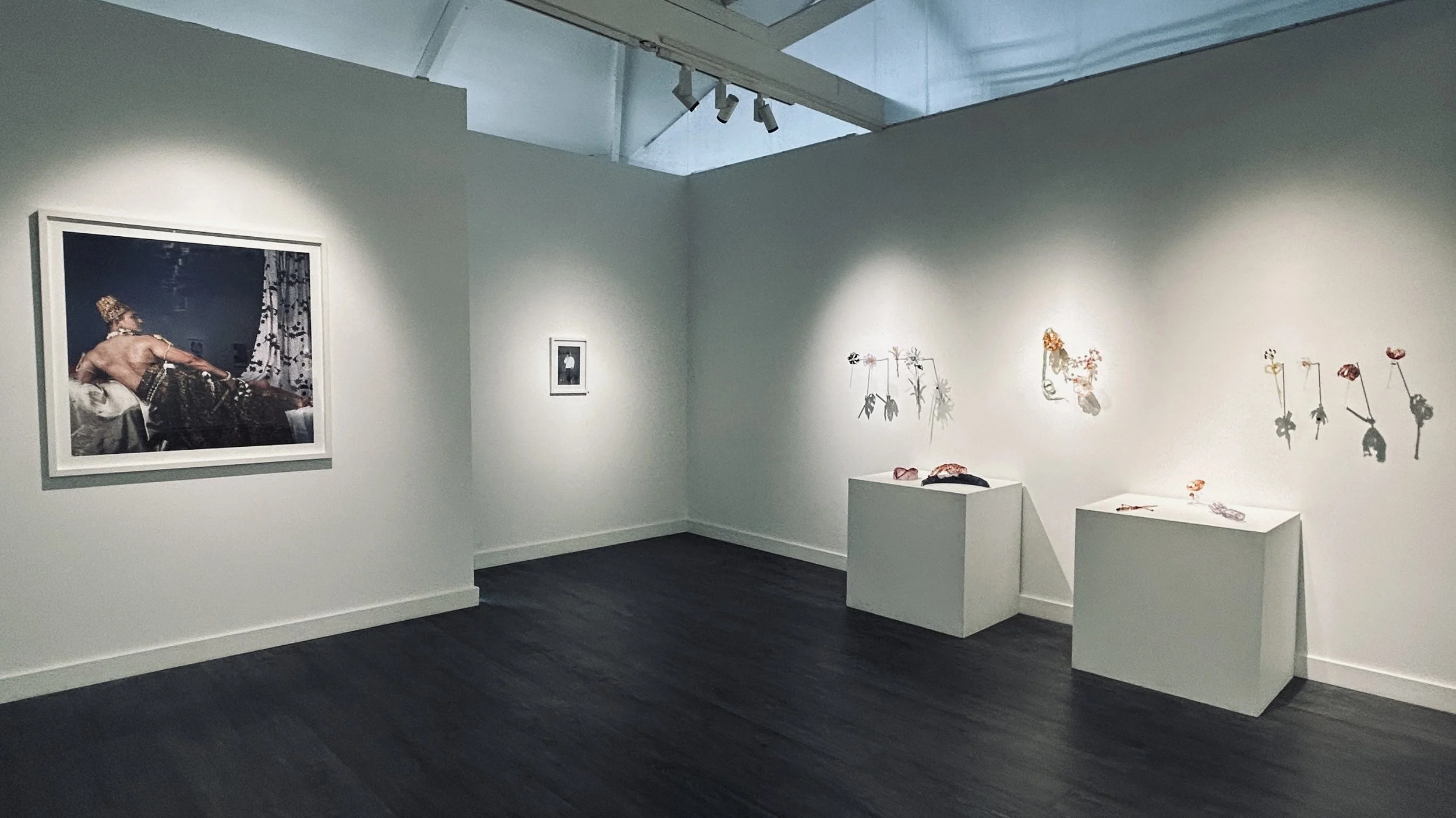 An art gallery with white walls displaying artwork, including a prominent portrait of a person wearing traditional attire and headdress, and floral-themed art pieces arranged on the walls and pedestals.