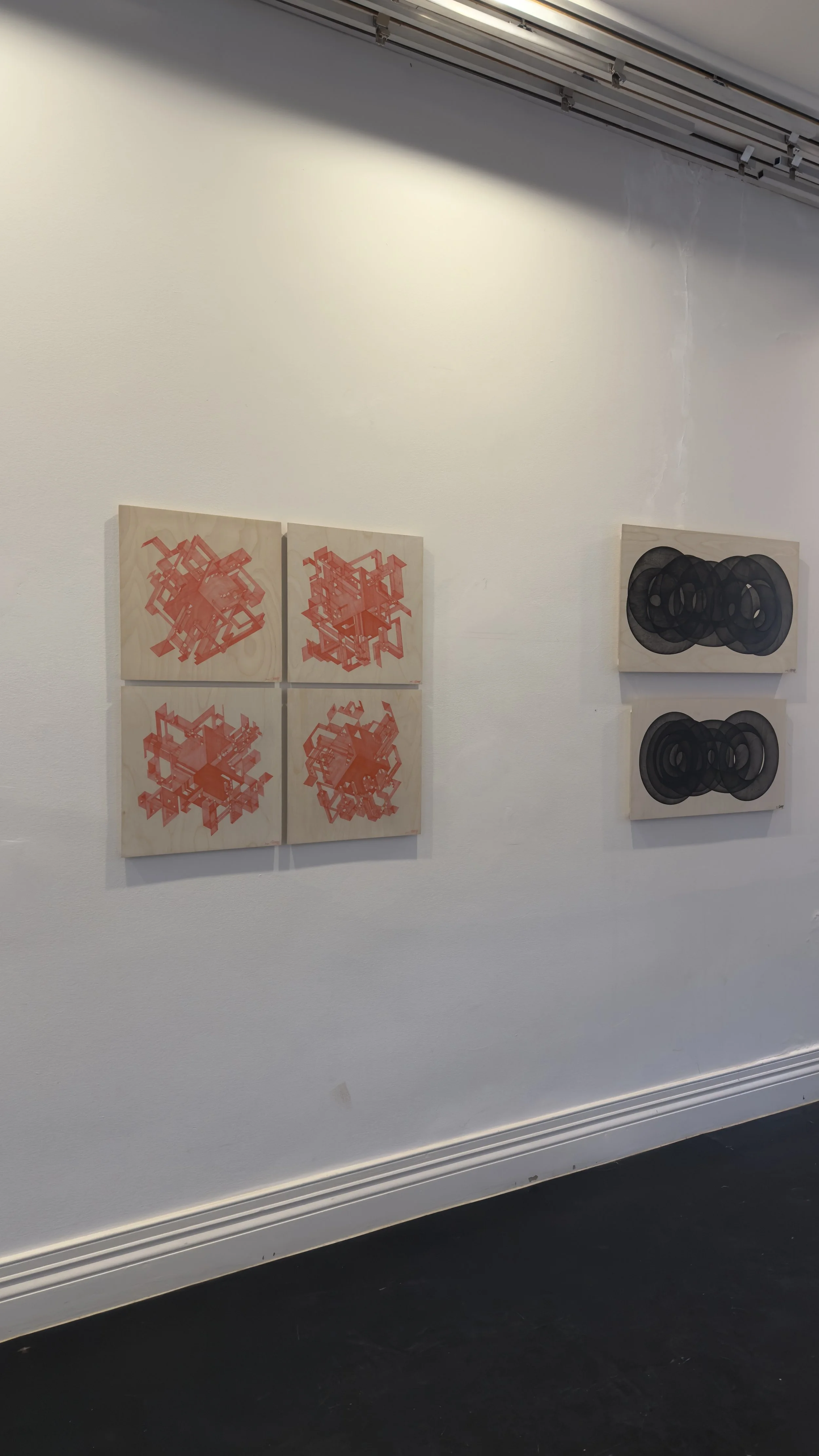 Four square abstract artworks with red geometric designs on the left and two square black and gray abstract designs on the right, all mounted on a white gallery wall.