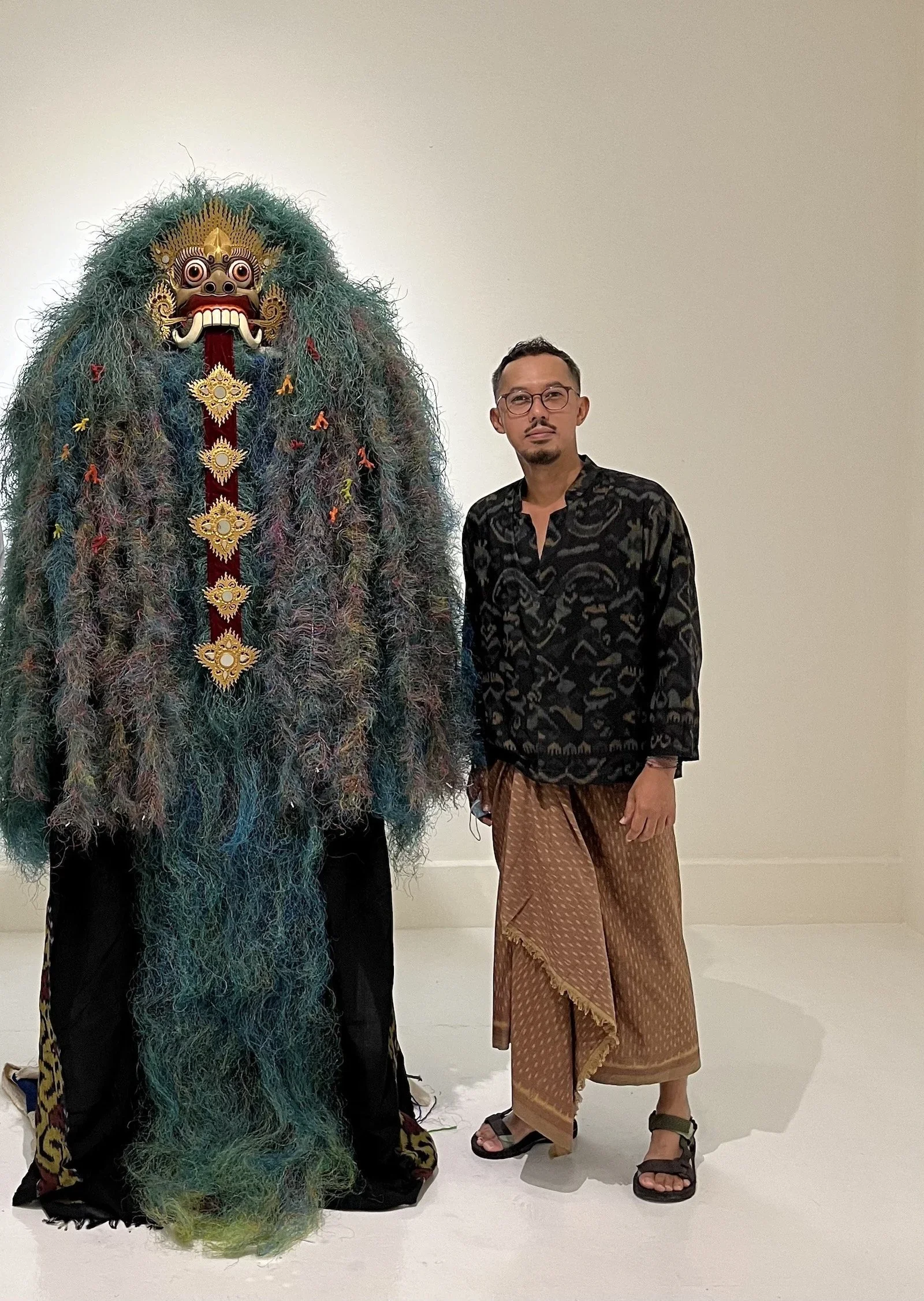 A person standing next to a traditional lion dance costume with a fierce lion mask and colorful, shaggy fur, in front of a plain white wall.