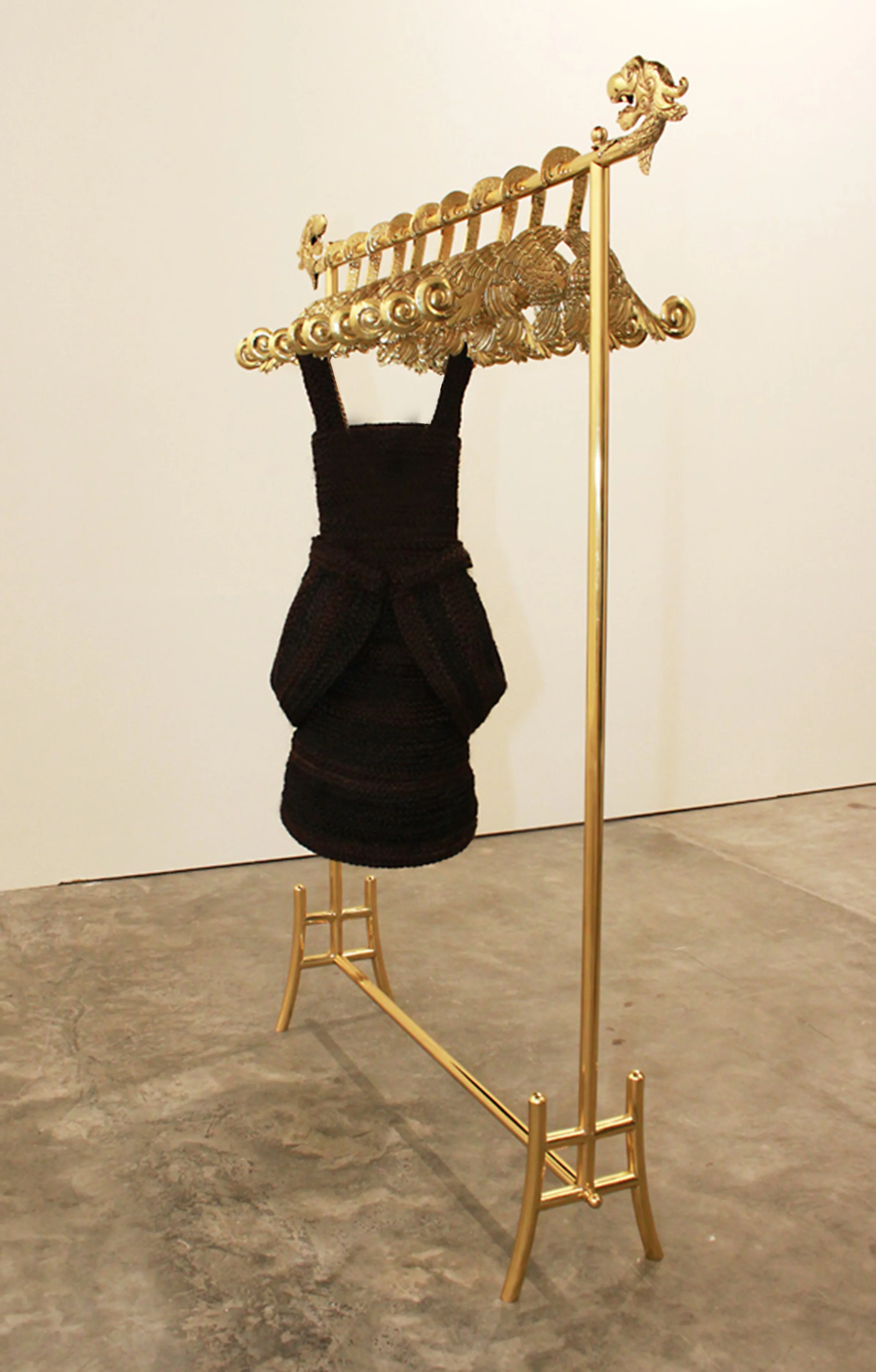 An art installation features a black crochet dress displayed on a stand that resembles a shoulder sculpture, topped with a gold ornate decorative element with intricate patterns, and supported by a gold stand resembling a clothing rack. The exhibit i