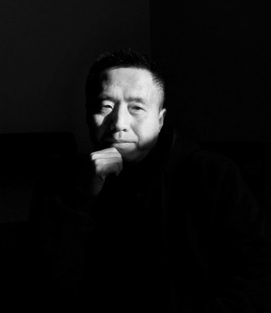 Black and white photo of a man with short hair, partially illuminated, resting his chin on his hand, in a dark environment.