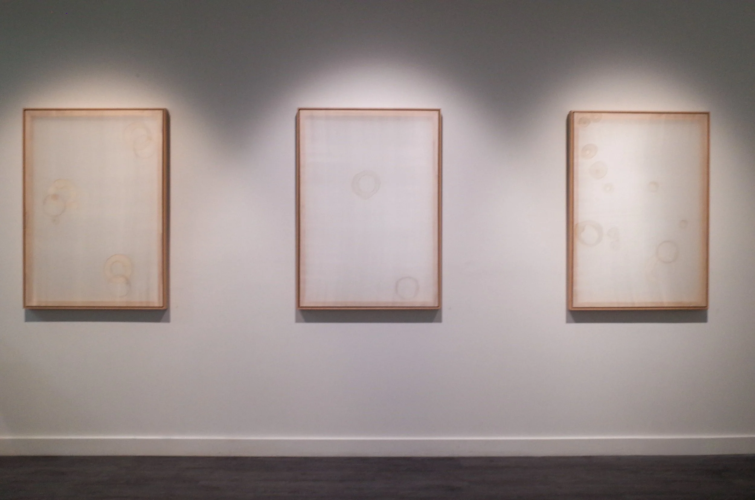 Three large white art pieces with faint circular patterns displayed on a wall in a gallery.