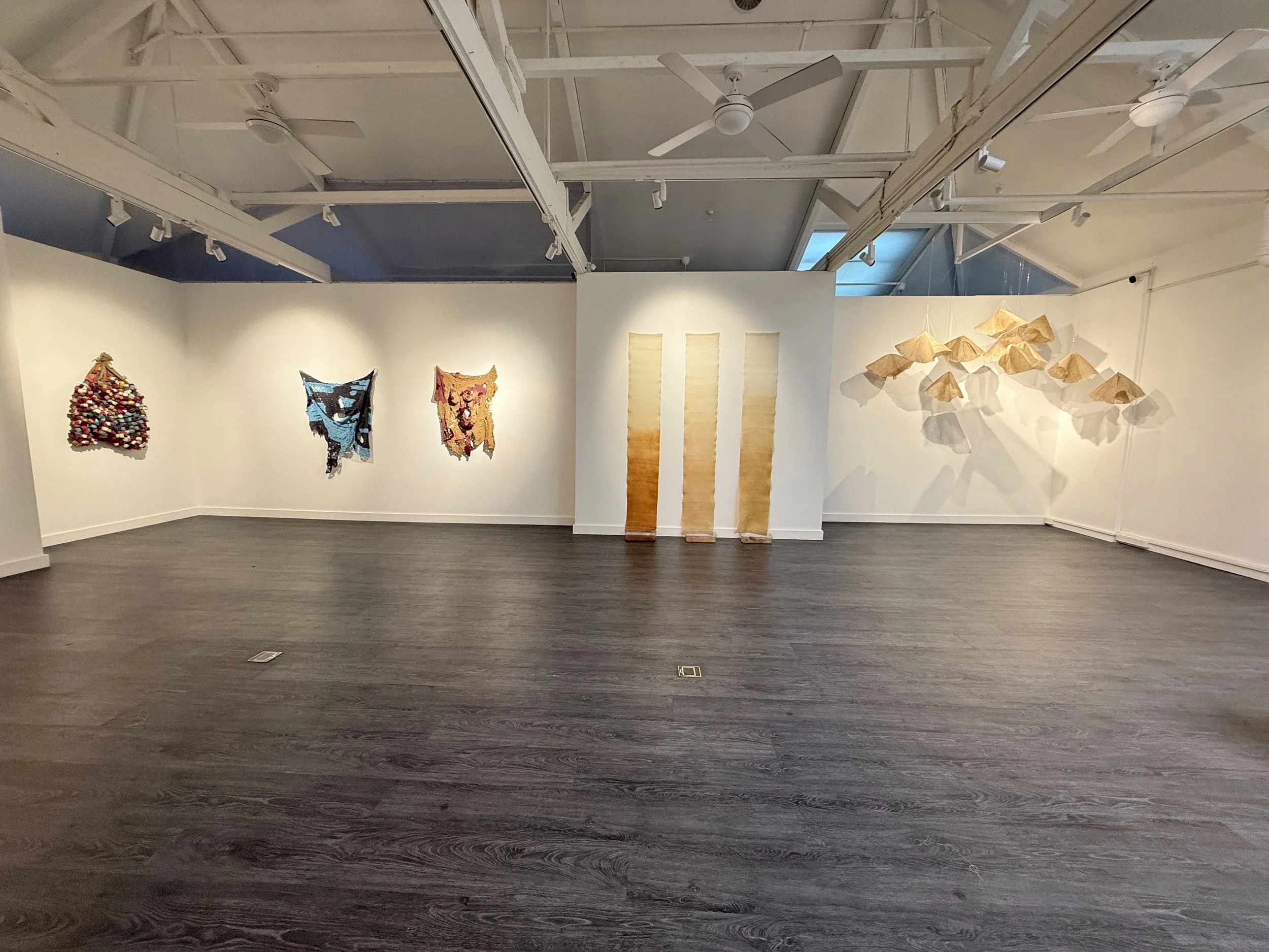 Art exhibition with woven textile wall hangings and paper sculptures in a gallery.