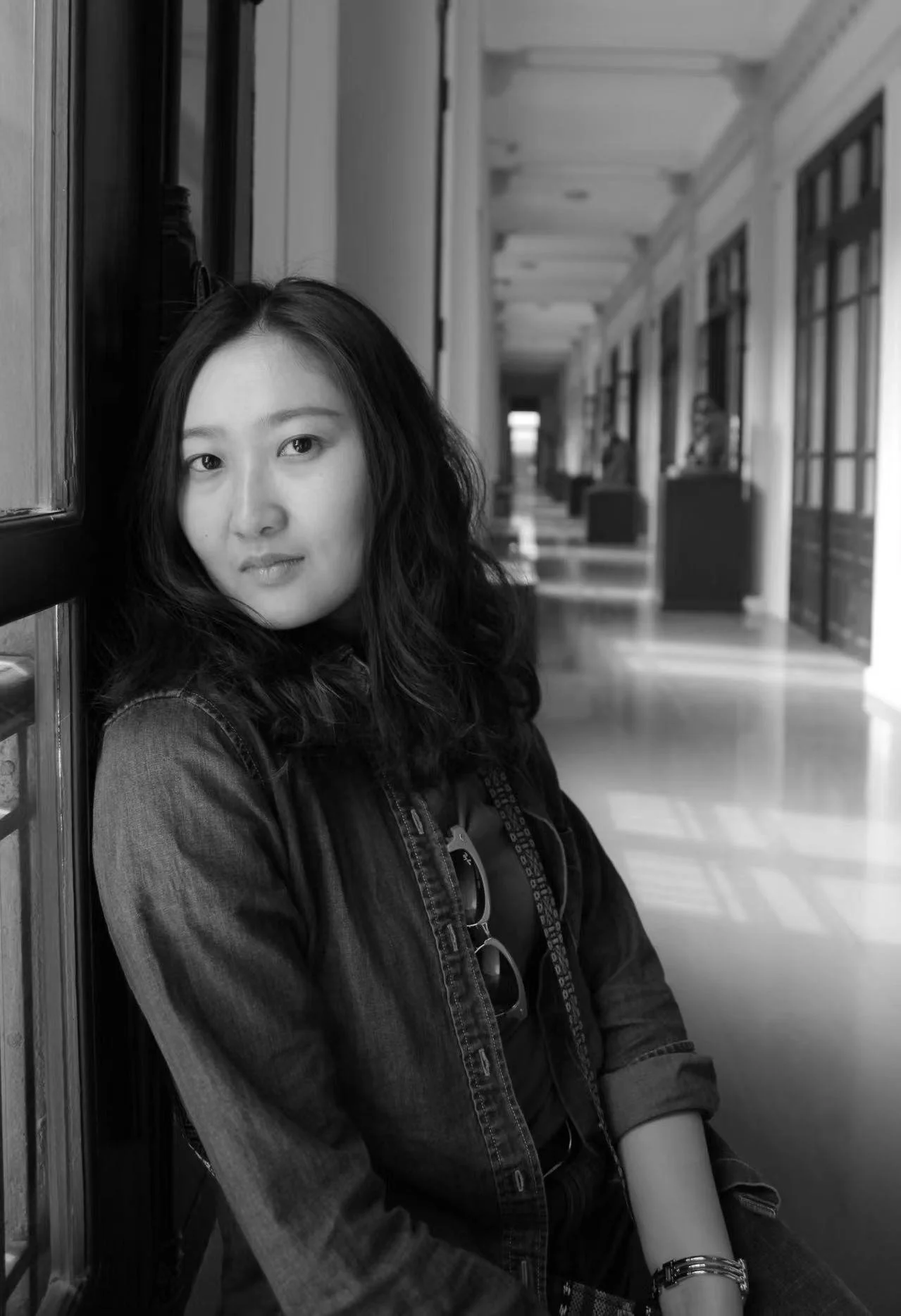 Black and white photo of a woman with wavy hair sitting against the wall of a corridor with windows and doors, looking at the camera.