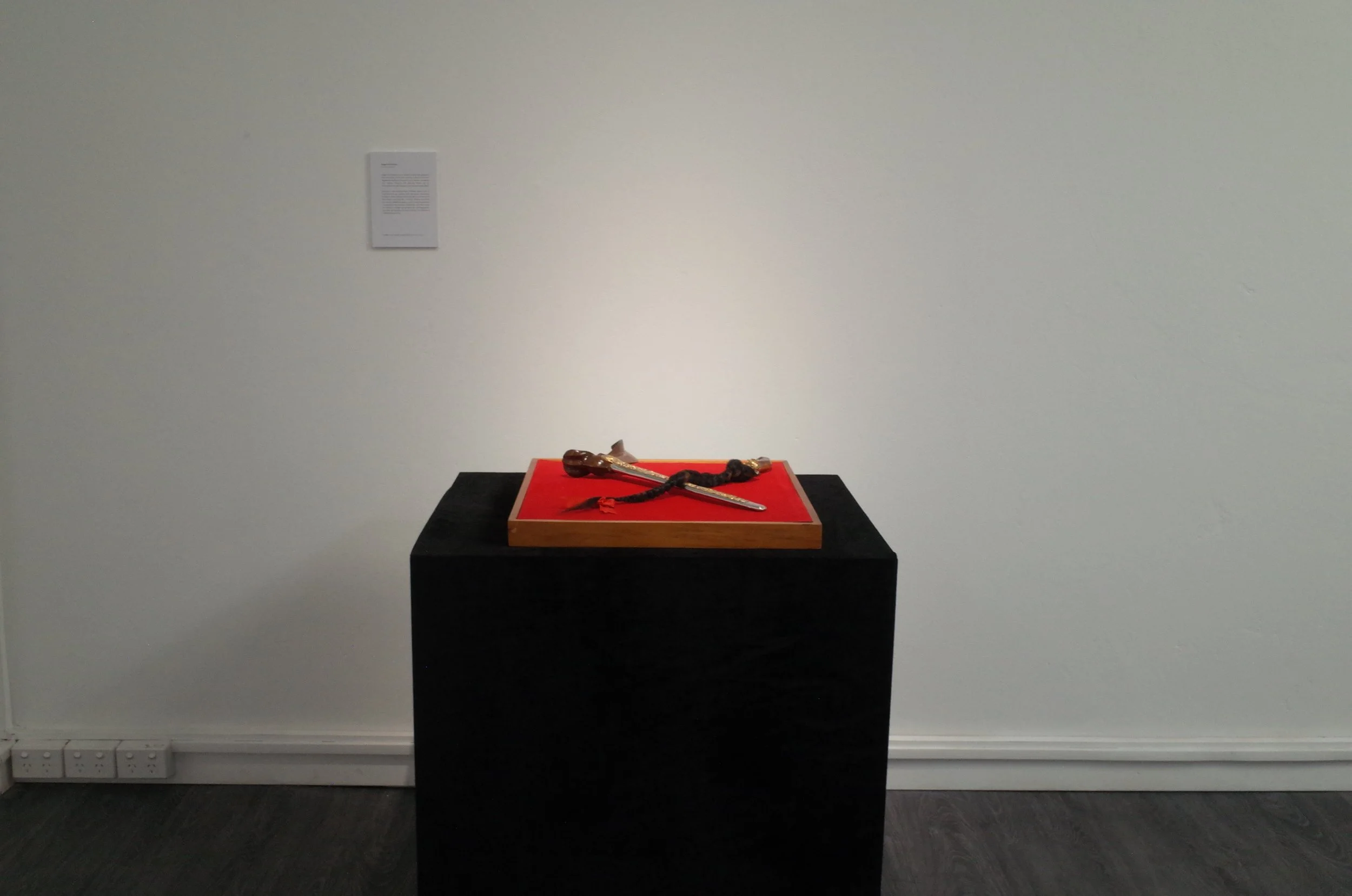 An art display featuring a carved wooden sculpture of a snake wrapped around a small object, placed on a red cloth on a black pedestal, against a plain white wall with a small informational plaque.