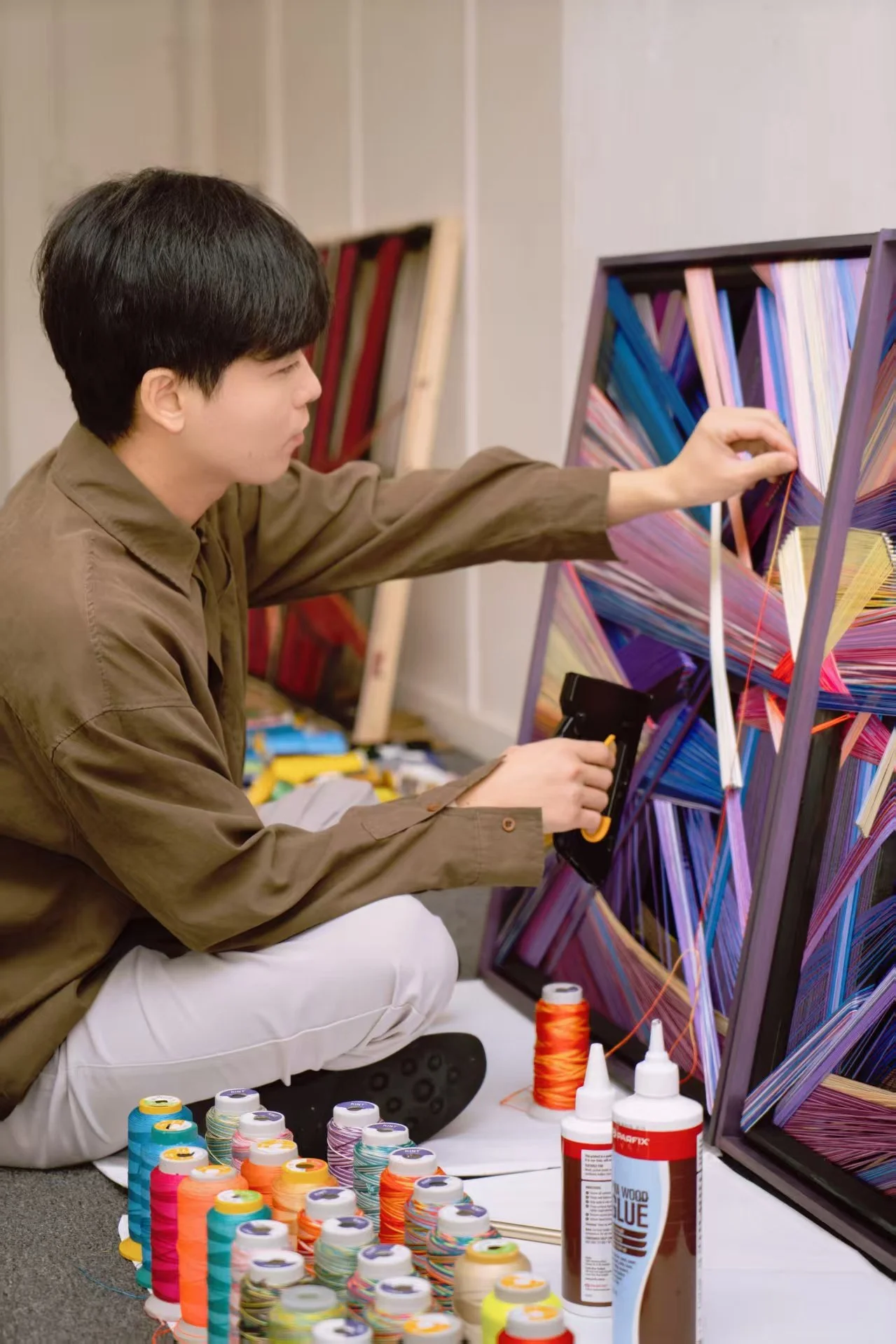 A person is sitting on the floor working on a colorful geometric art piece on a large picture frame. There are multiple spools of colorful thread, bottles of glue, and other art supplies around them.