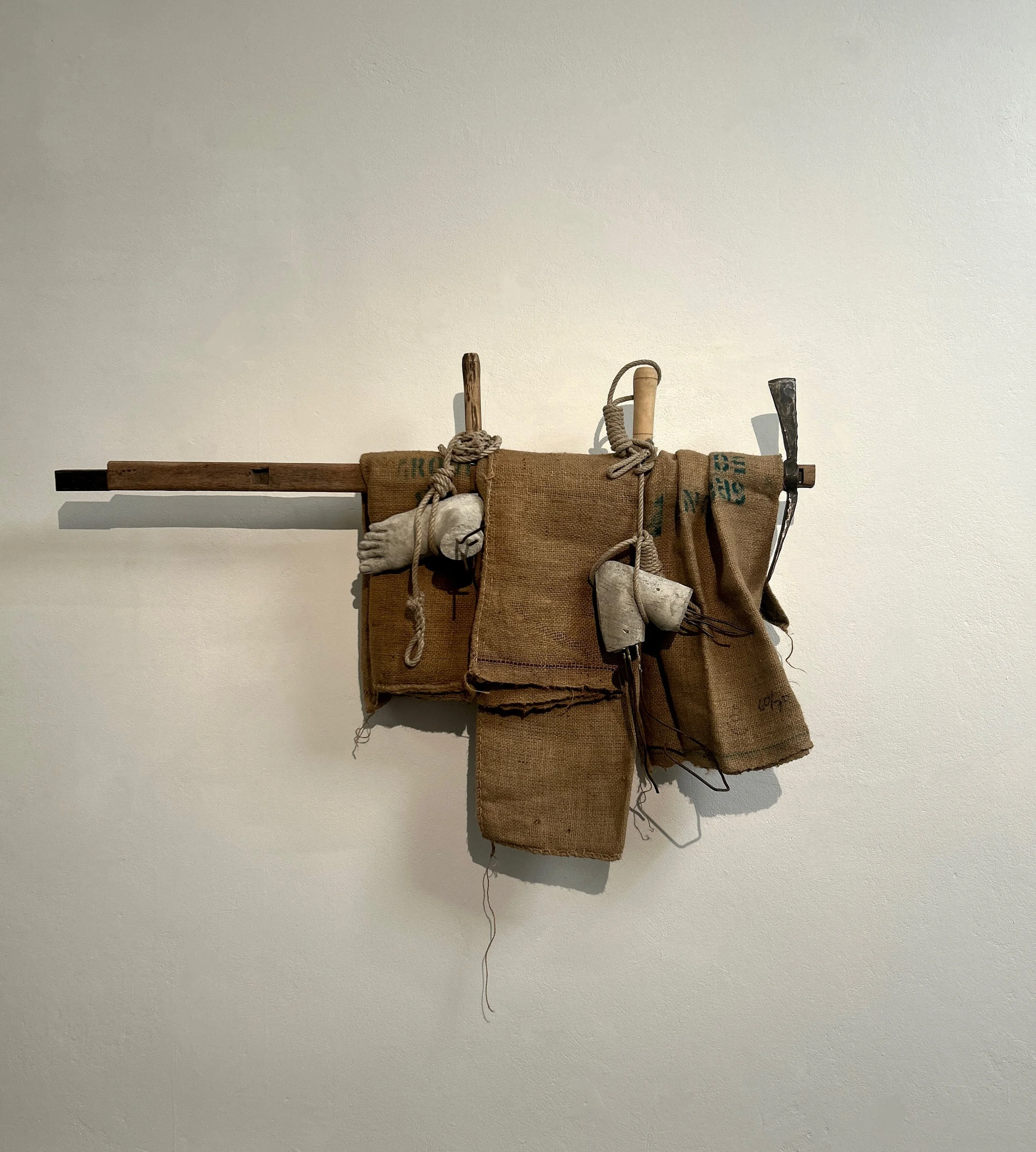 An art installation featuring a horizontal wooden board with various objects attached, including canvas bags, rope, metallic tools, and a foot-like sculpture, all mounted on a white wall.