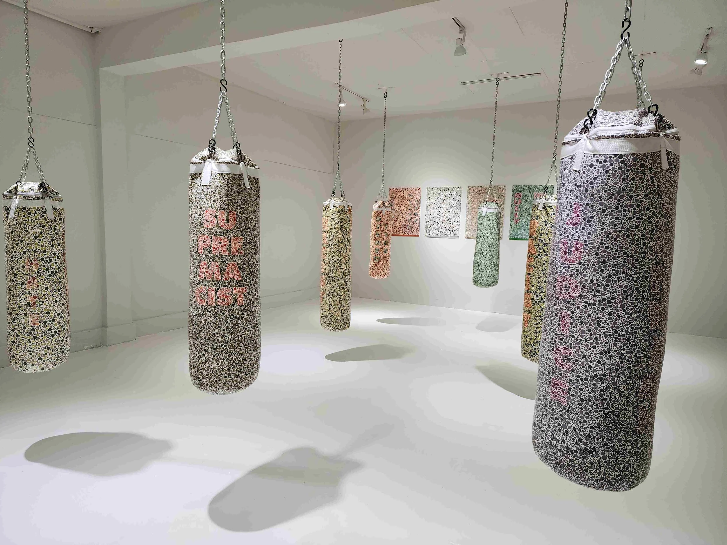 Art installation of punching bags covered in a dotted pattern with colorful text, hanging from the ceiling in a white gallery space.