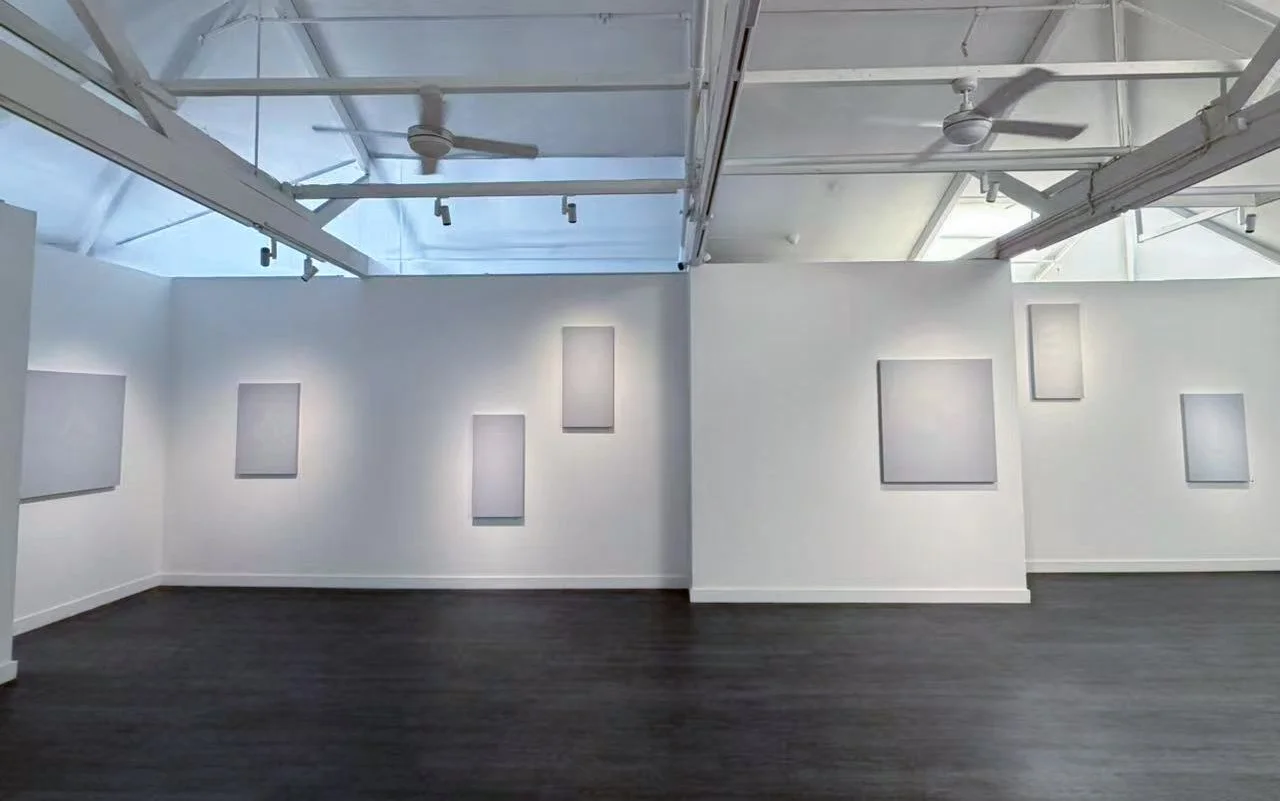 An art gallery with white walls and dark hardwood flooring, featuring several blank, framed canvases on the walls. The ceiling has skylights and is equipped with ceiling fans and track lighting.