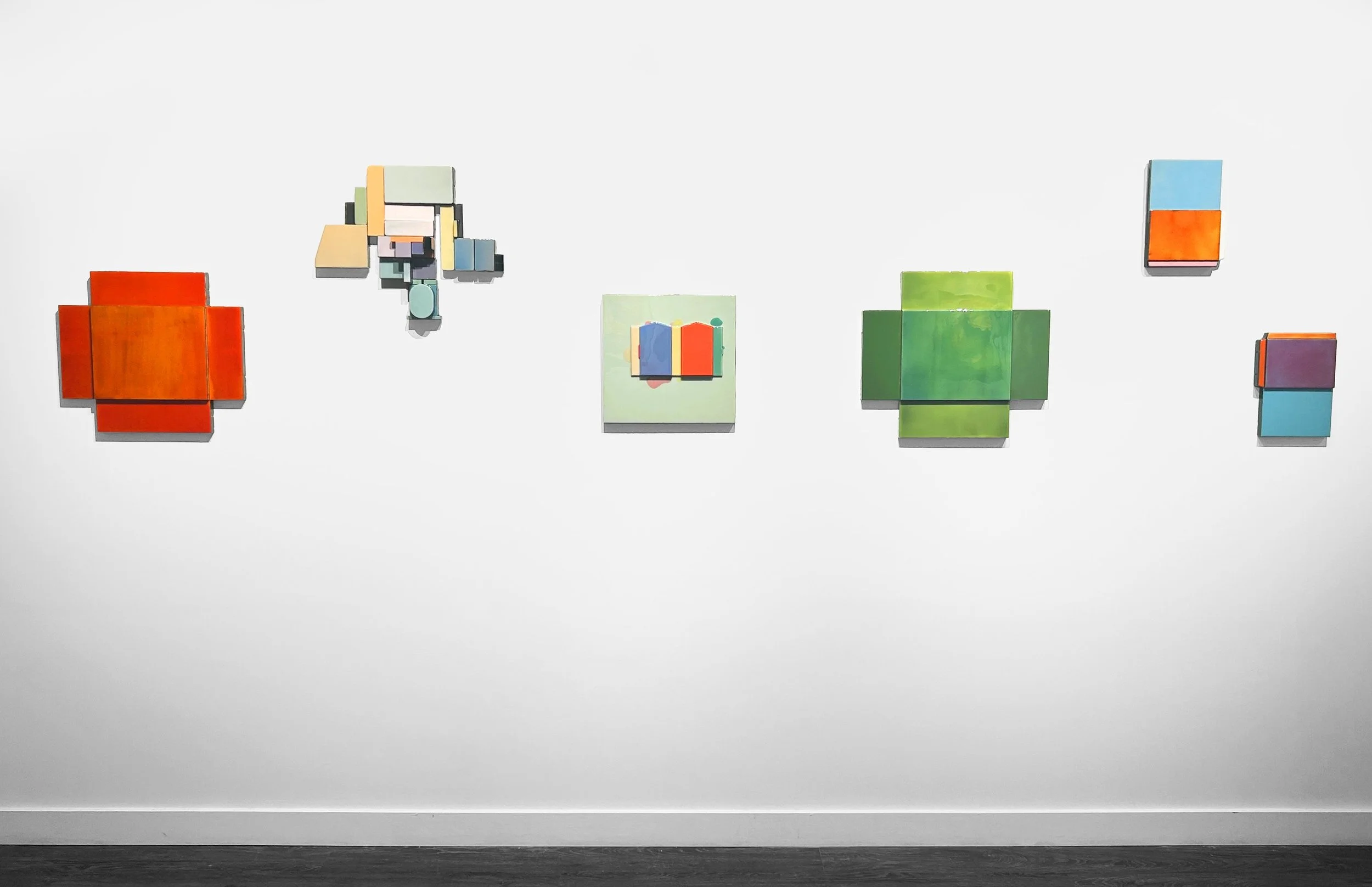 Minimalist art installation of colorful geometrical shapes on a white gallery wall, arranged in a linear pattern.