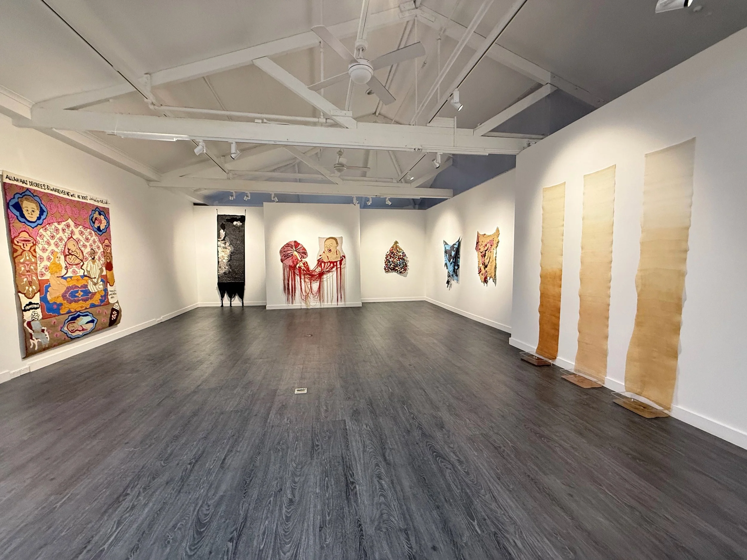 An art gallery with white walls displaying colorful textile art pieces, including a large portrait, abstract shapes, and hanging woven fabrics, with a gray hardwood floor and ceiling lights.
