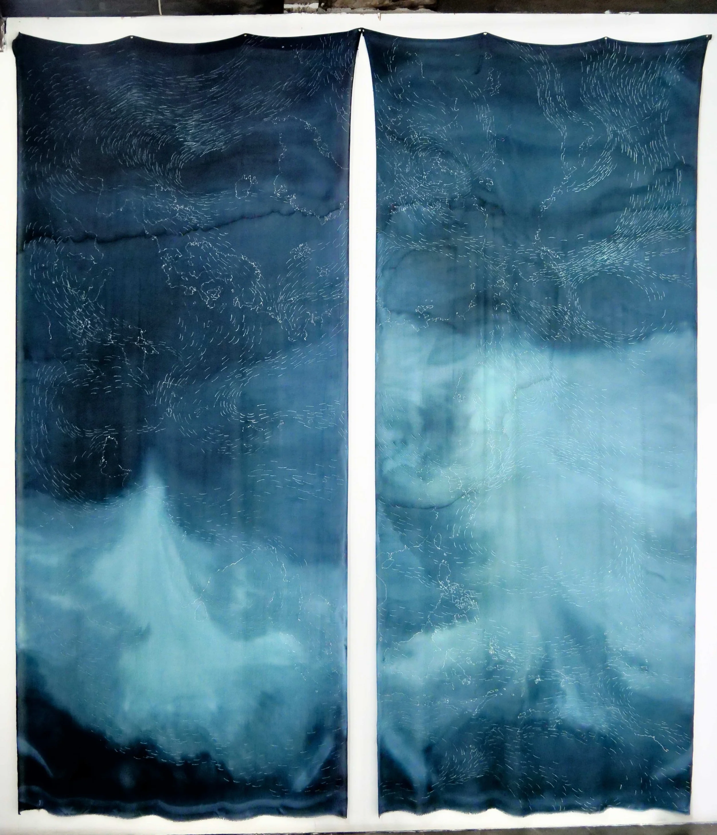 Two large blue fabric curtains with abstract white line designs resembling weather patterns or star trails, hung side by side on a white wall.