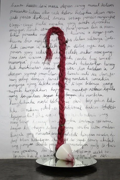 A handmade craft consisting of a heart-shaped base with a red yarn braid extending upward, attached to a white heart-shaped top. The background features handwritten text in black ink on white paper.