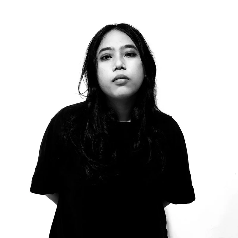 Black and white portrait of a young woman with long dark hair, wearing a dark shirt, standing against a plain white background.