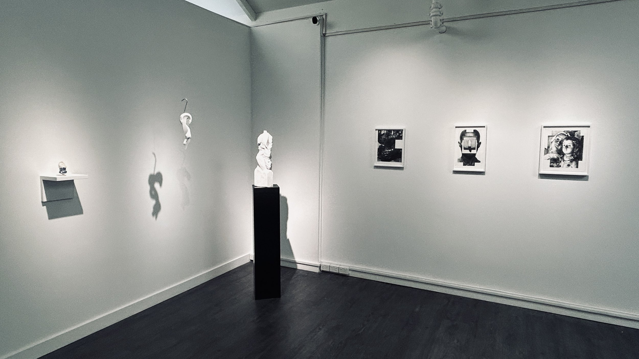An art gallery with three framed photographs on a white wall, a sculpture on a pedestal, and a small sculpture on a wall-mounted shelf. Shadows of the sculptures are cast on the wall.