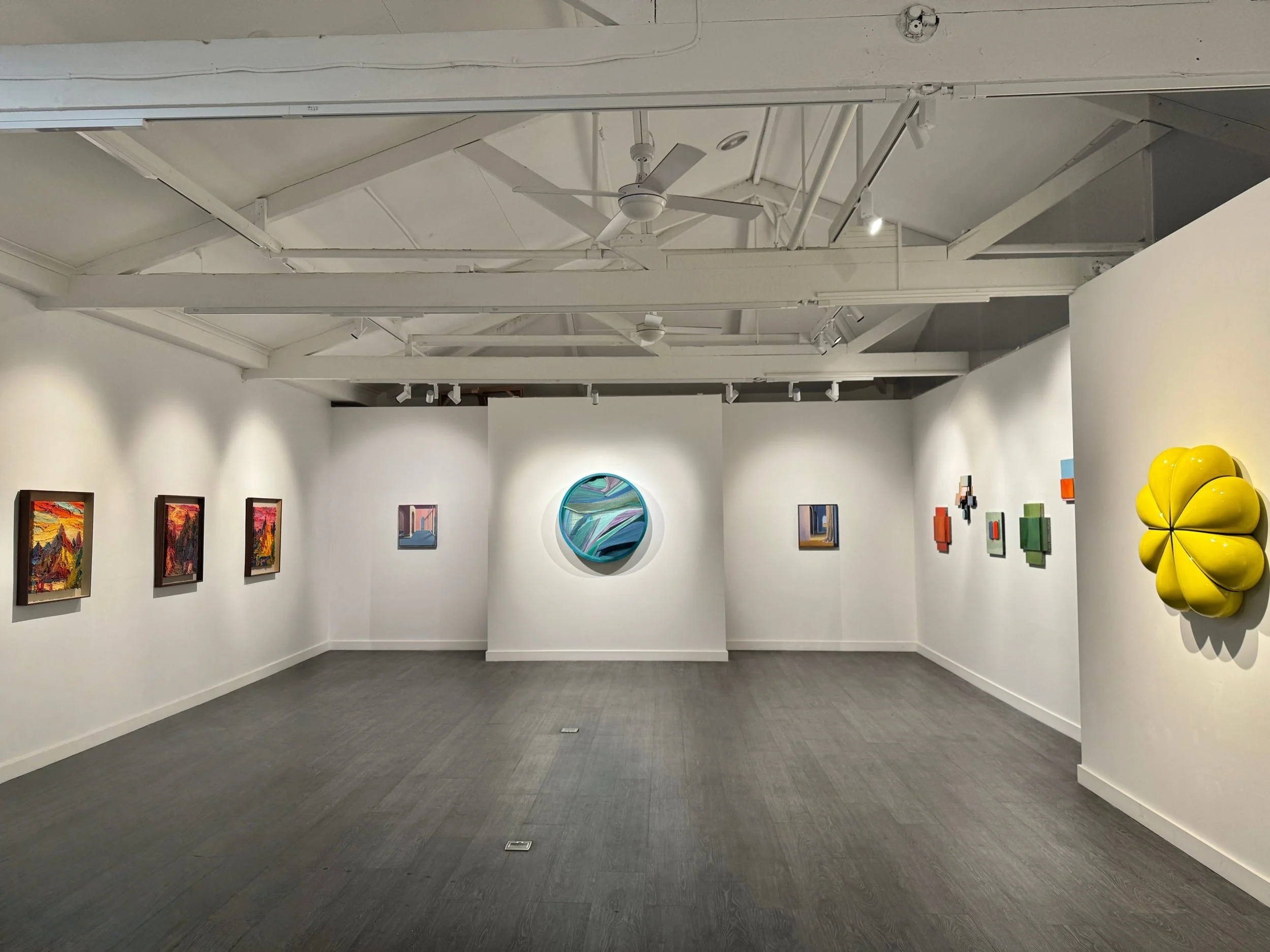 Interior of an art gallery with paintings on the walls, a ceiling fan, and sculptures including a yellow flower-shaped piece.