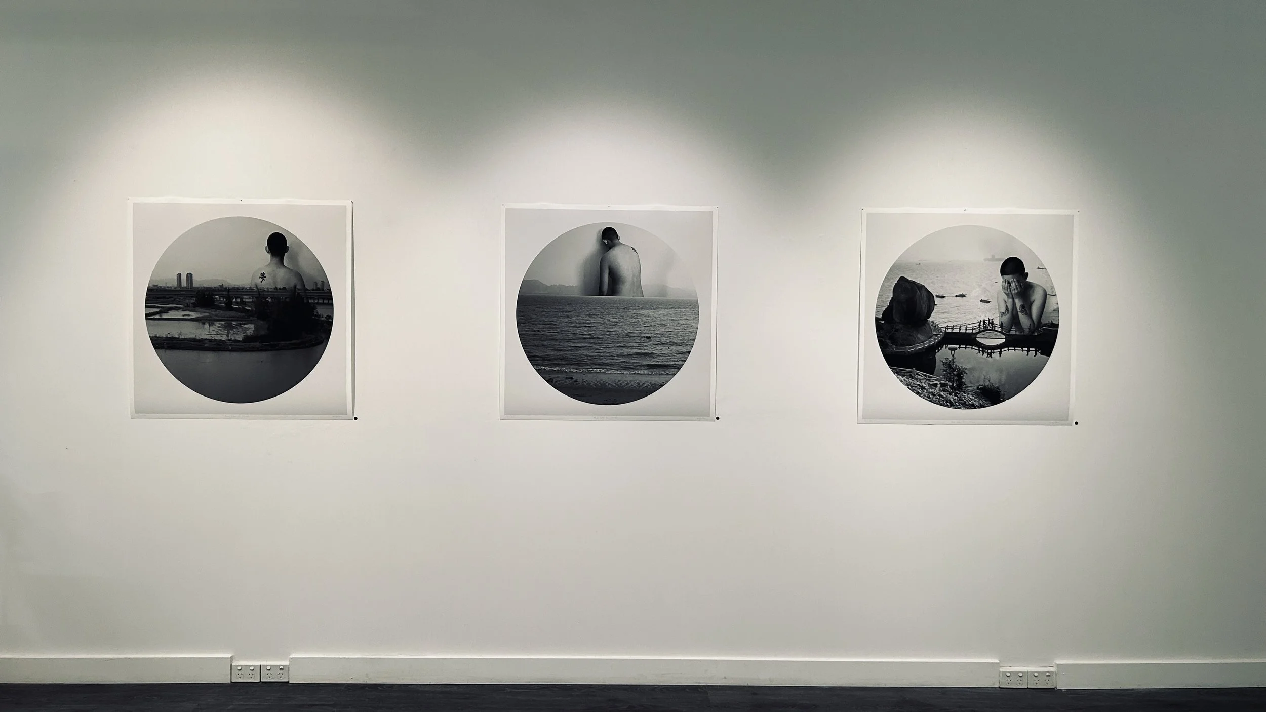 Three black-and-white photographs of women, each framed in a circular border, hung on a gallery wall. The first shows a woman with her back to the camera, overlooking a cityscape. The second depicts a woman with her back to the camera, facing the sea