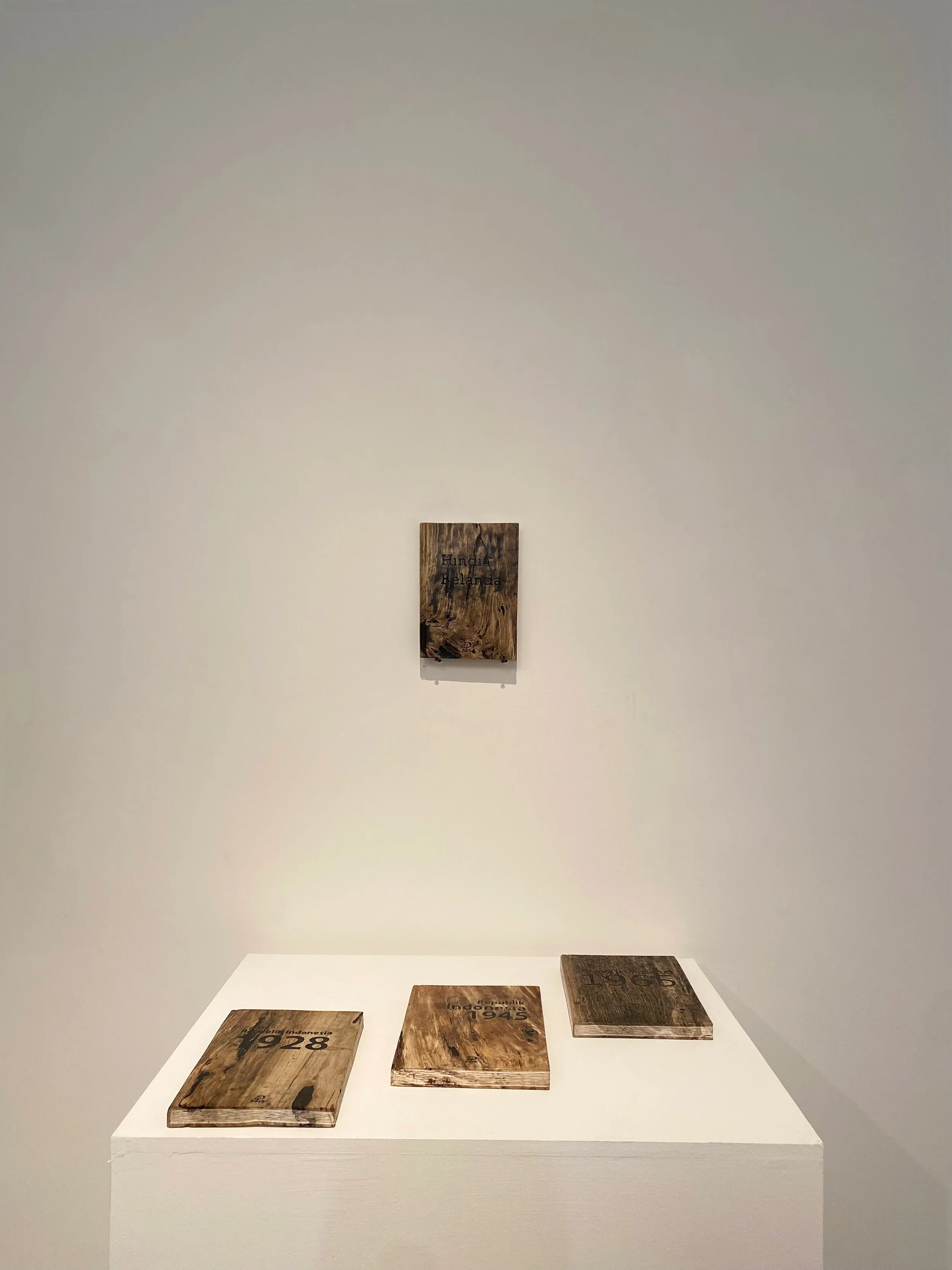 An art exhibit with five wooden plaques placed on a white pedestal. The plaques have various dates and inscriptions, including 1928, 1945, and 2015.