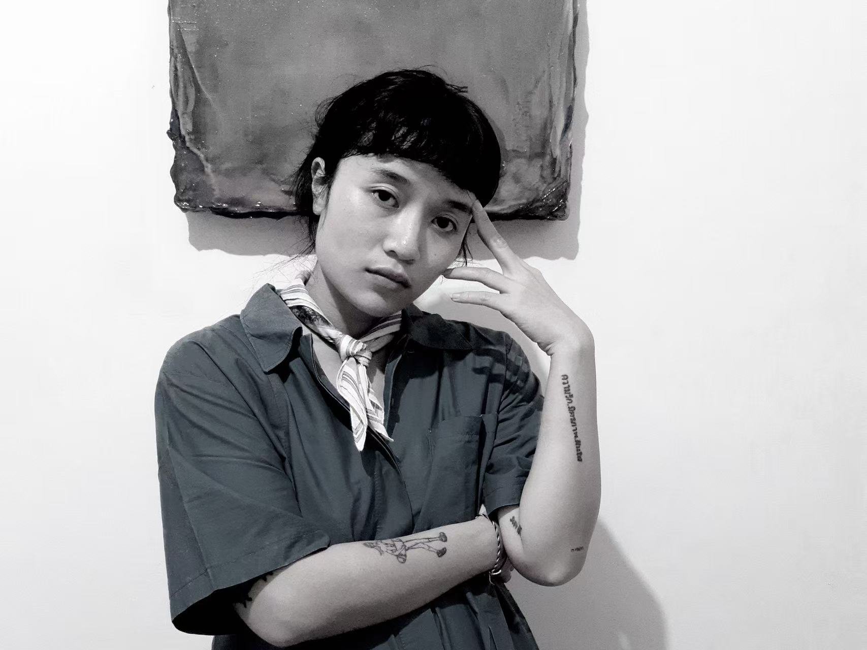 A black and white photo of a woman with short dark hair, wearing a button-up shirt and a striped necktie, standing against a plain wall with a textured painting behind her. She has tattoos on her arms, and her right hand is touching her forehead while her left arm is crossed over her body.