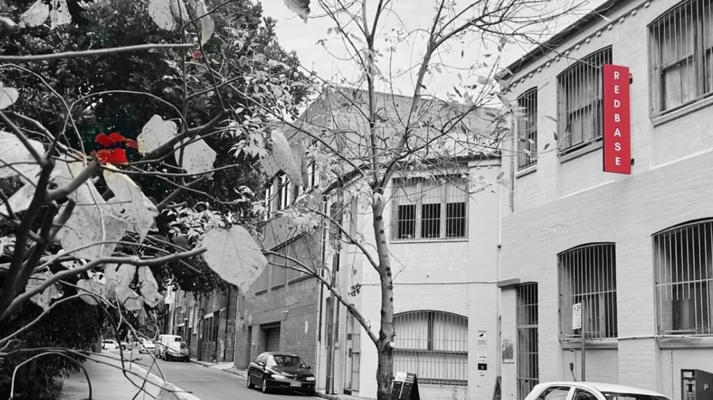 Street scene with cars parked along the side, buildings with barred windows, a red vertical sign that reads 'REDBASE,' and trees with sparse leaves, some snow on the branches.