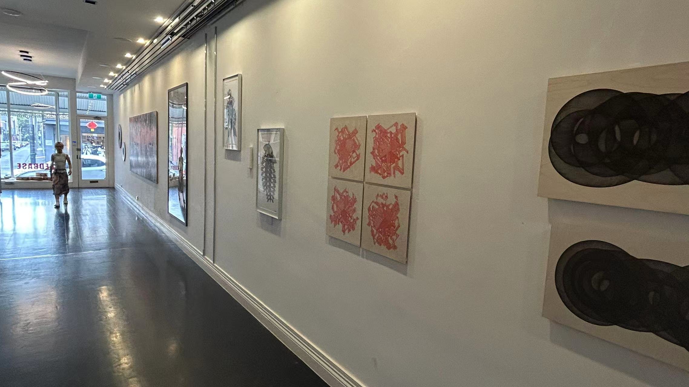 An art gallery with artwork displayed on the white walls, a person walking near the entrance, and large windows letting in natural light.