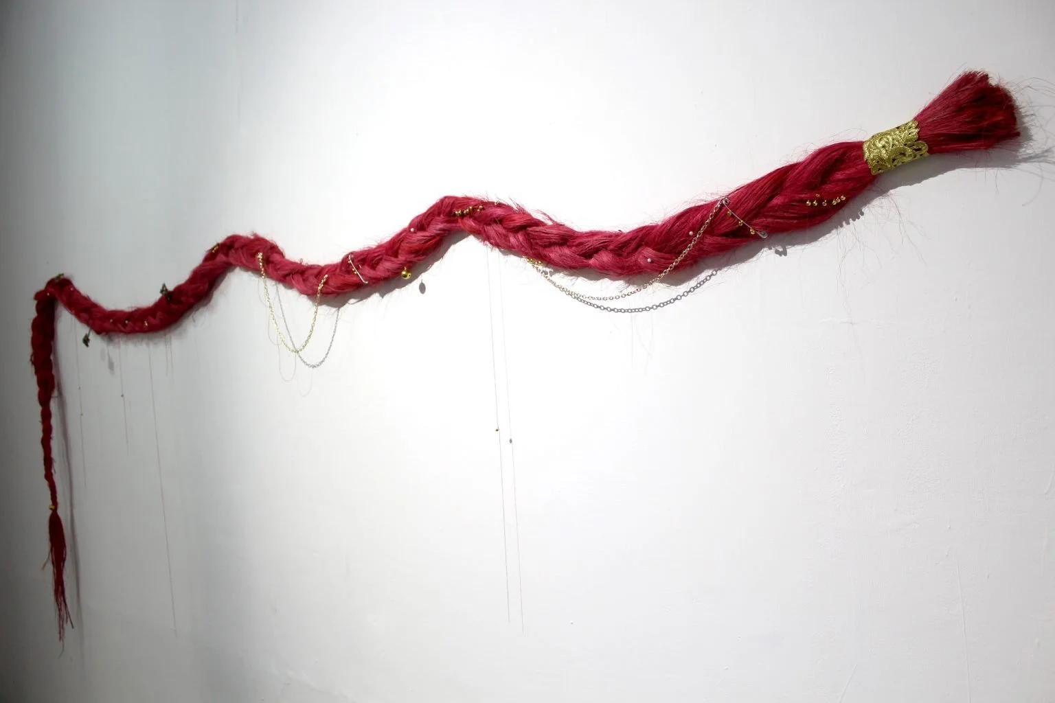An art installation on a white wall featuring a long, twisted, red hair-like sculpture decorated with small gold chains, beads, and a gold clasp at one end.