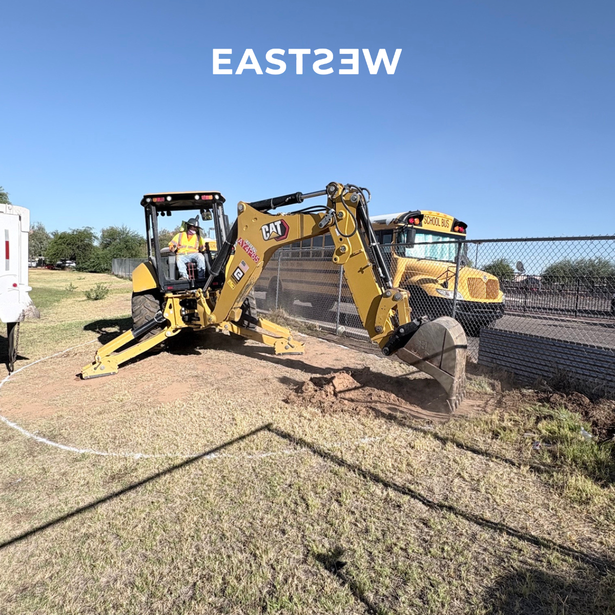 A construction worker operates a yellow CAT backhoe excavator on a grassy area, near a school bus in the background, with a blue sky overhead.