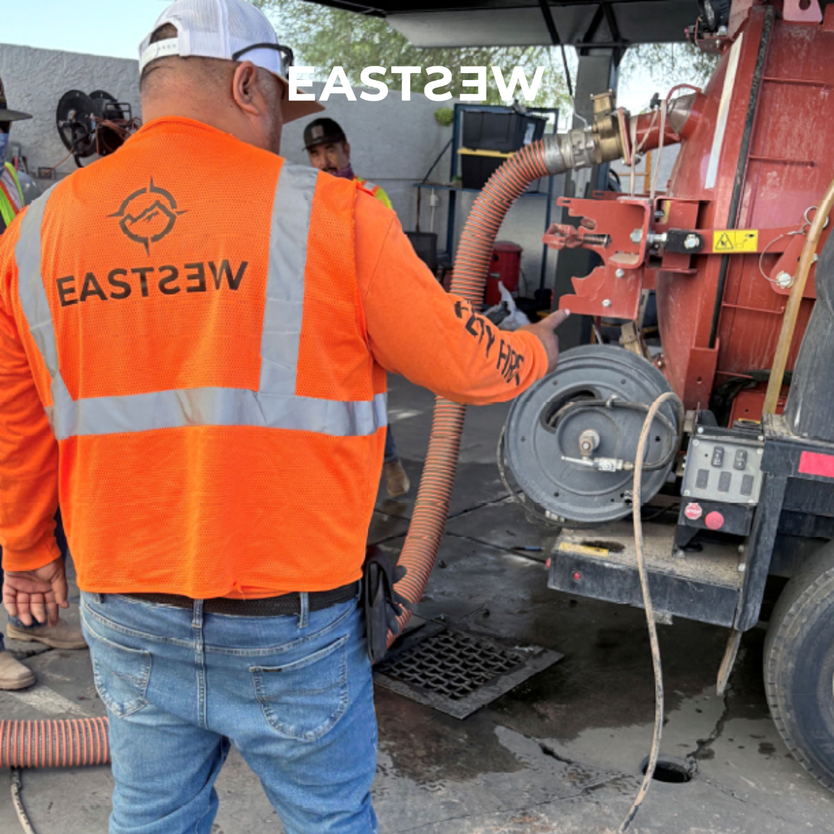 Worker in an orange safety vest and jeans operating industrial equipment outdoors, with several other workers nearby.