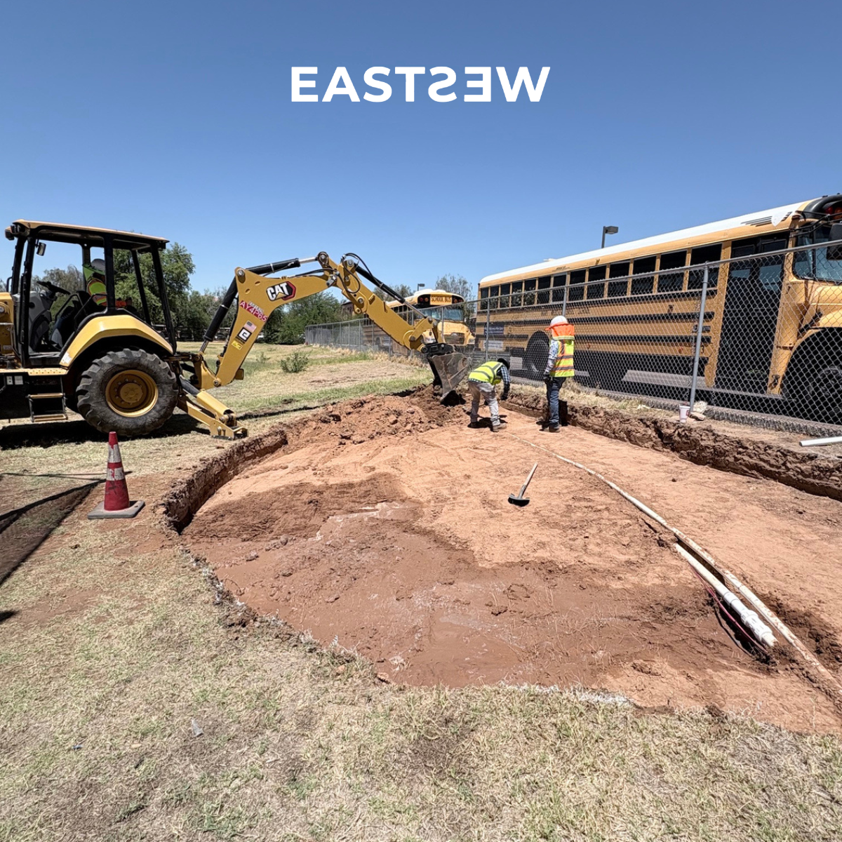 Construction site with excavator digging a trench, workers in safety vests and helmets, school bus and construction vehicles nearby, sign indicating east direction.