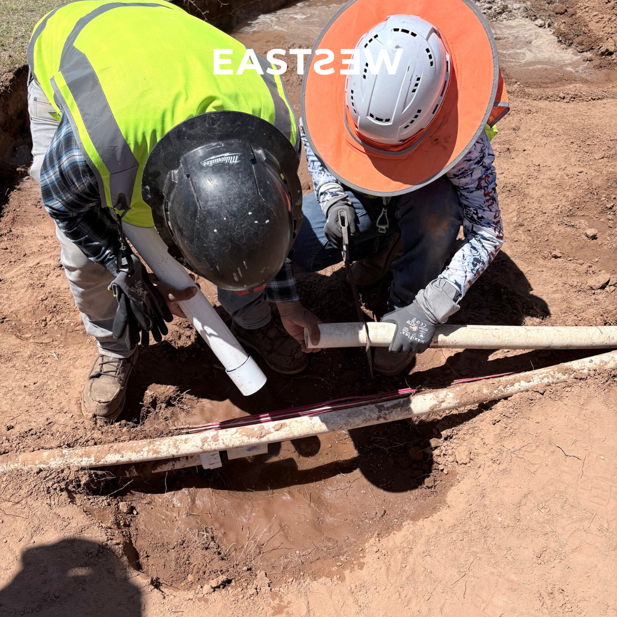 Two construction workers wearing safety helmets, gloves, and reflective vests are working together on a pipe underground in a dirt area, possibly installing or repairing it.