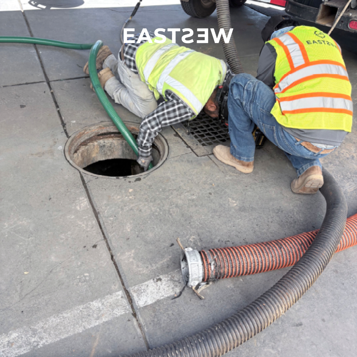 Two workers in safety vests working near an open storm drain on a city sidewalk, with hoses connected to equipment.