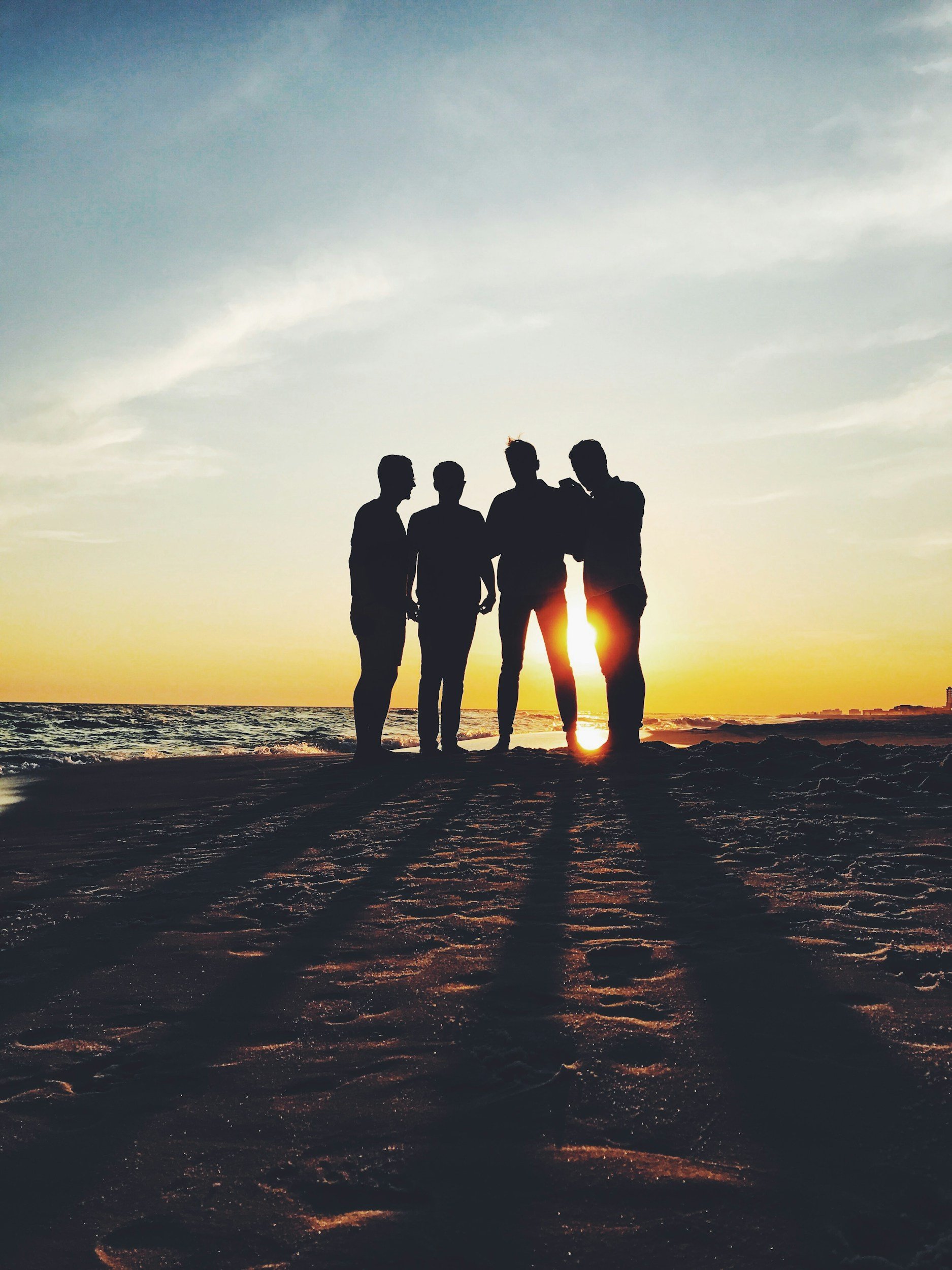 Four people are standing on the beach at sunset, silhouettes against the sky, with the sun low near the horizon.