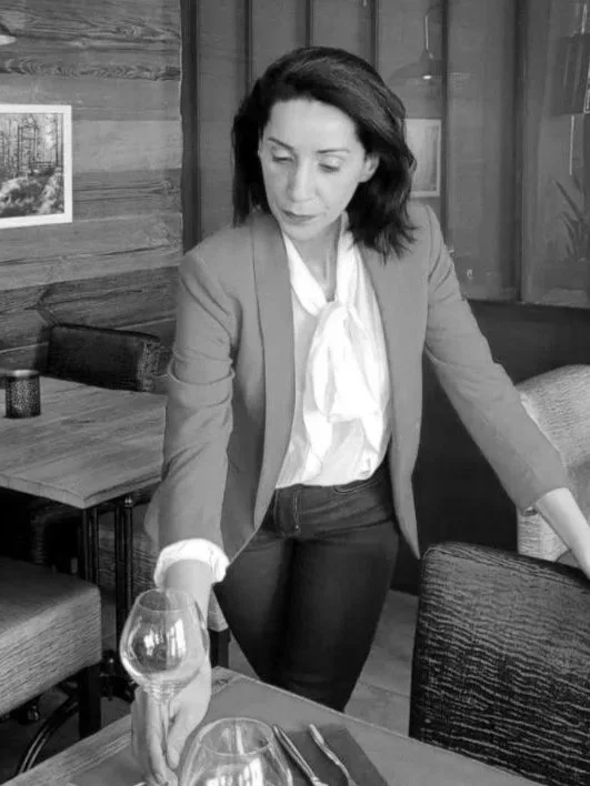 Femme debout à une table dans un restaurant, ajustant une nappe ou un verre, portant une veste et une chemise blanche, en noir et blanc.
