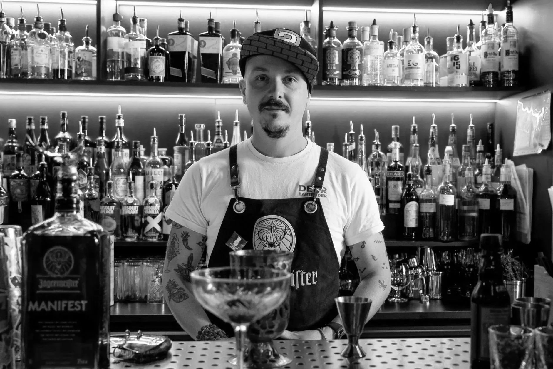 Barman devant un bar avec de nombreuses bouteilles d'alcool sur des étagères, portant un t-shirt blanc, un tablier noir et un chapeau à carreaux.